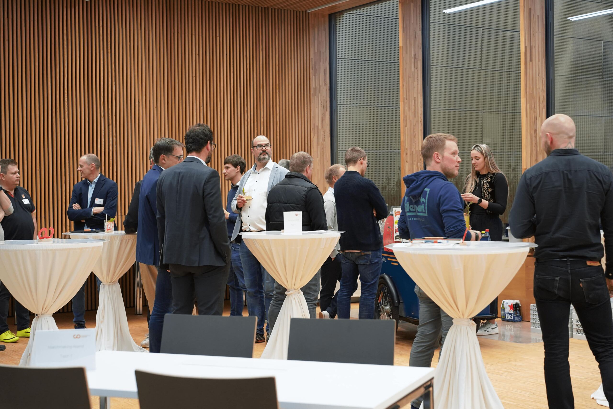 Eine Gruppe von Menschen unterhält sich bei einer Networking-Veranstaltung. Sie stehen in einem modernen Raum an hohen, runden Tischen mit weißen Tischdecken. Einige halten Getränke in der Hand, und rechts ist eine Frau in Schwarz zu sehen. Der Raum hat große Fenster und Holzvertäfelungen.