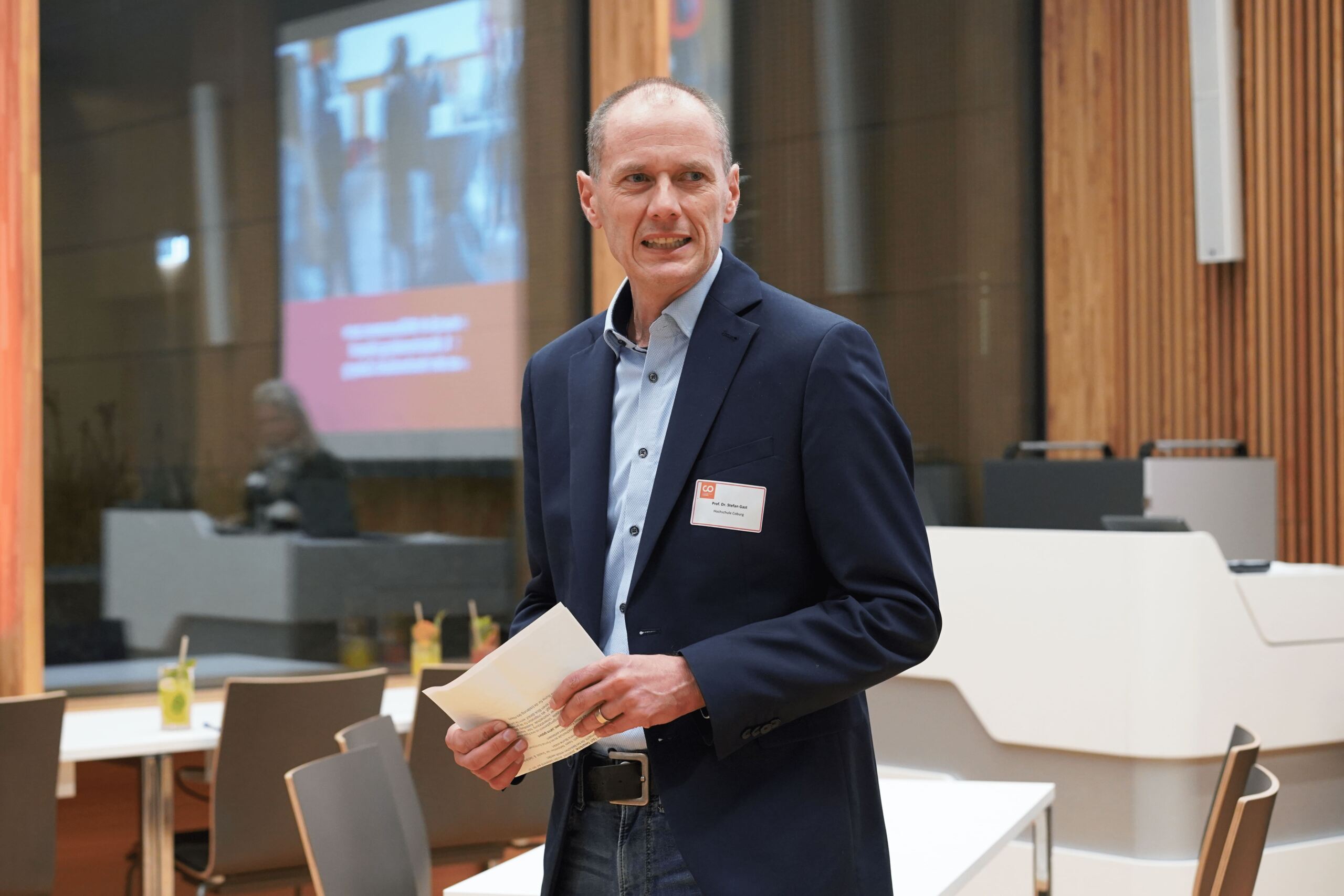 Ein Mann in einem marineblauen Blazer und einem blauen Hemd steht drinnen, hält Papiere in der Hand und spricht. Hinter ihm stehen Konferenztische und ein großer Bildschirm, auf dem ein unscharfes Bild und Text angezeigt werden. Die Umgebung scheint ein moderner Konferenz- oder Besprechungsraum zu sein.