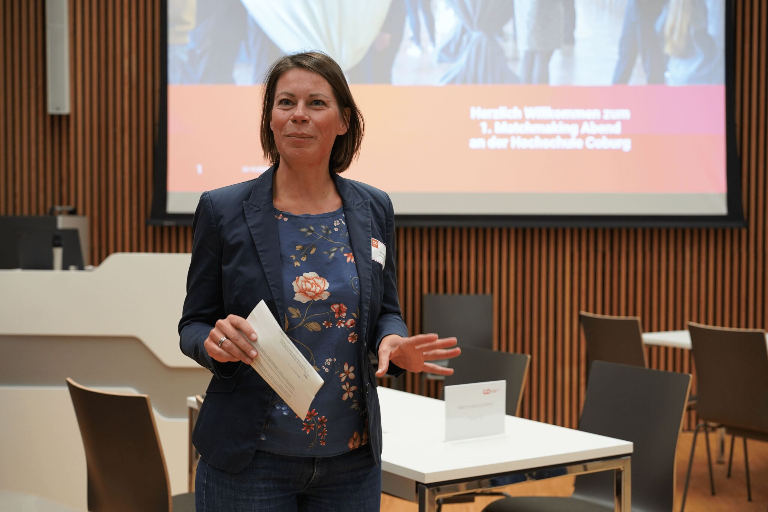 Eine Frau steht drinnen und hält ein Stück Papier in der Hand. Sie trägt einen marineblauen Blazer über einem geblümten Hemd und Jeans. Hinter ihr ist ein Präsentationsbildschirm mit Text und mehrere Tische und Stühle zu sehen.