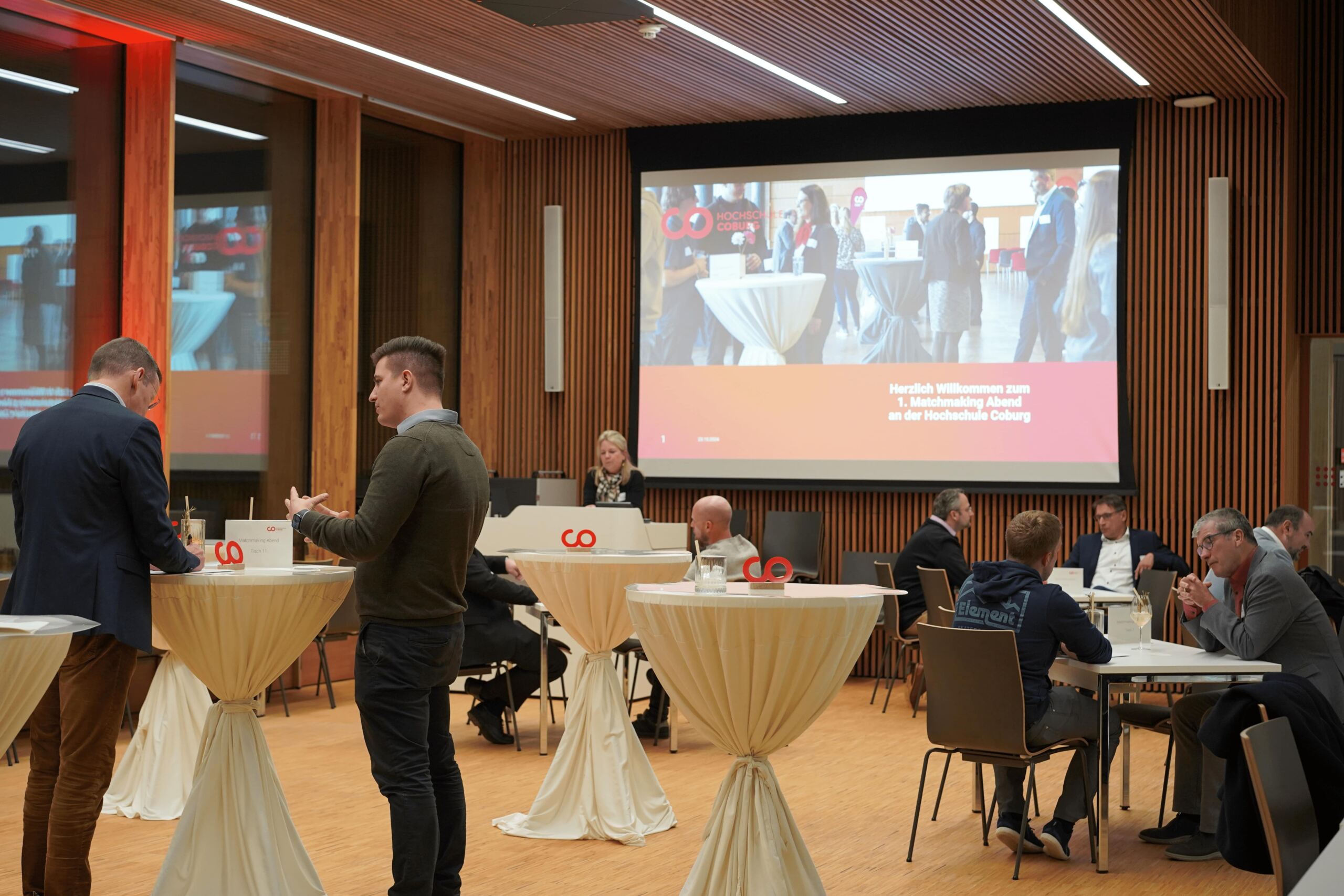 In einem modernen Konferenzraum mit hohen Tischen sind Menschen versammelt. Auf einer Projektionsleinwand läuft eine Präsentation. Die Teilnehmer unterhalten sich und knüpfen Kontakte.