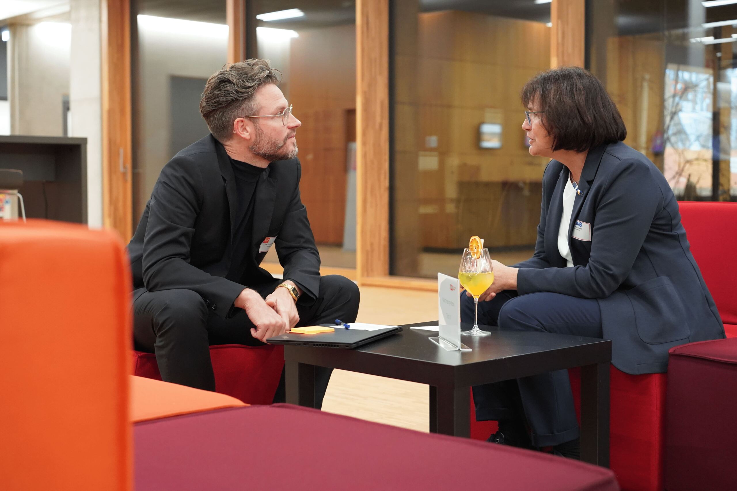 Ein Mann und eine Frau sitzen auf roten Sofas in einer Bürolounge und unterhalten sich. Auf dem Couchtisch zwischen ihnen stehen ein Notizbuch und ein Cocktailglas. Sie sind in Business-Kleidung gekleidet, im Hintergrund ist eine holzgetäfelte Wand zu sehen.