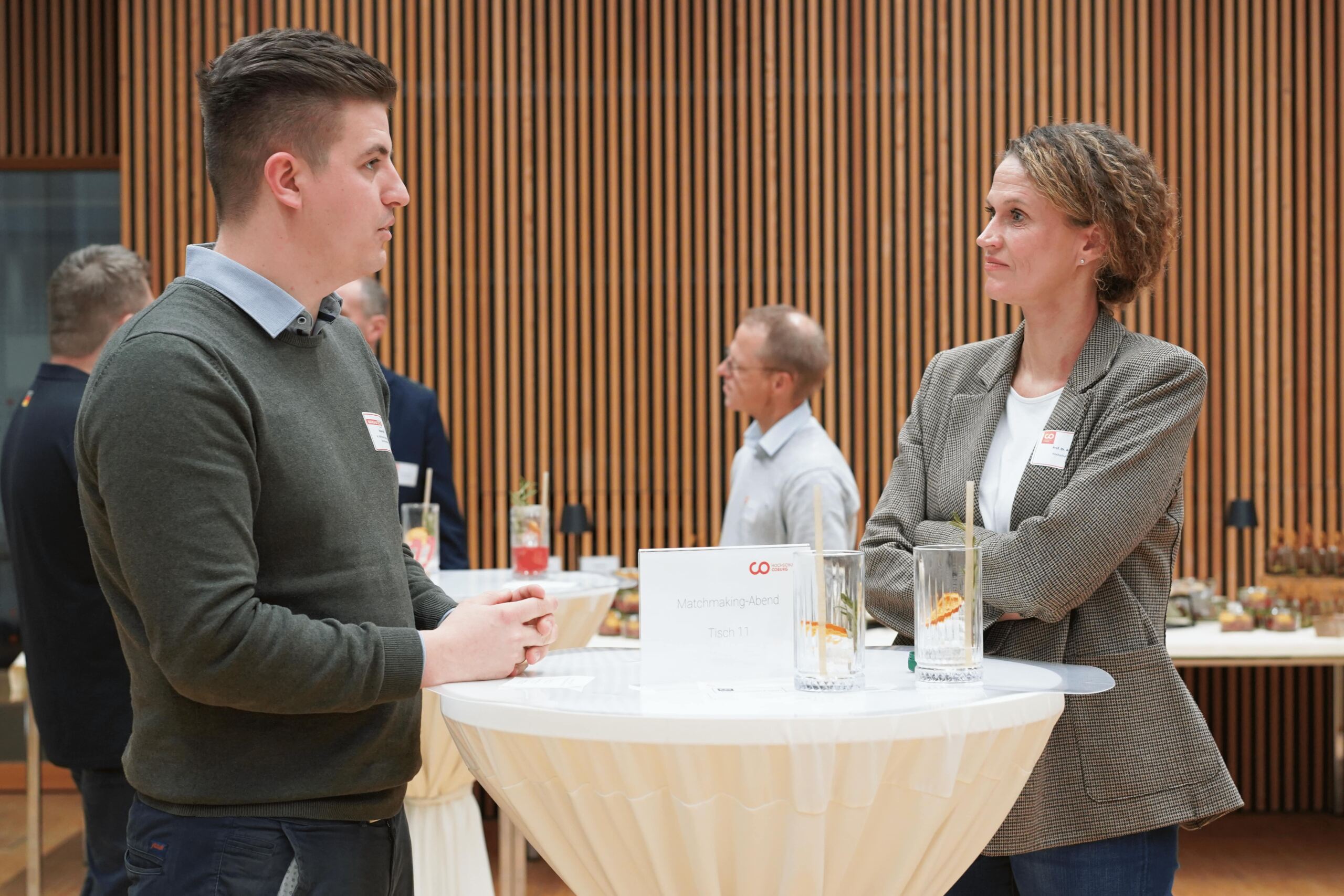 Zwei Personen stehen an einem runden Tisch und unterhalten sich. Auf dem Tisch stehen zwei leere Gläser und ein Schild. Im Hintergrund sind weitere Personen zu sehen, außerdem Holzwandpaneele und ein Buffettisch.