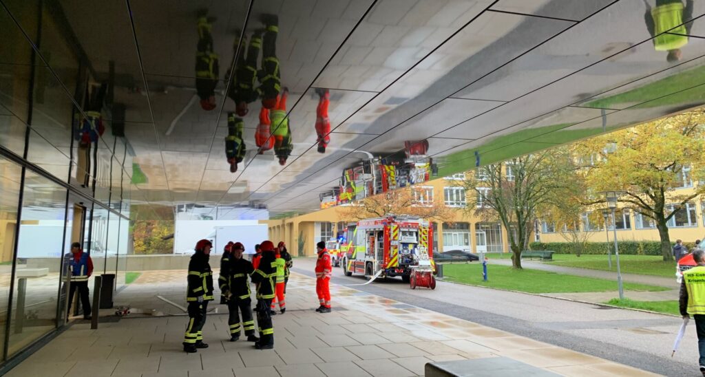 Feuerwehrmänner in Uniform stehen vor einem modernen Gebäude mit reflektierenden Glasscheiben. Ein Feuerwehrauto und mehrere Mitarbeiter sind zu sehen, im Hintergrund sind Bäume und eine Straße zu sehen. Reflexionen an der Decke zeigen gespiegelte Bilder der Szene darunter.