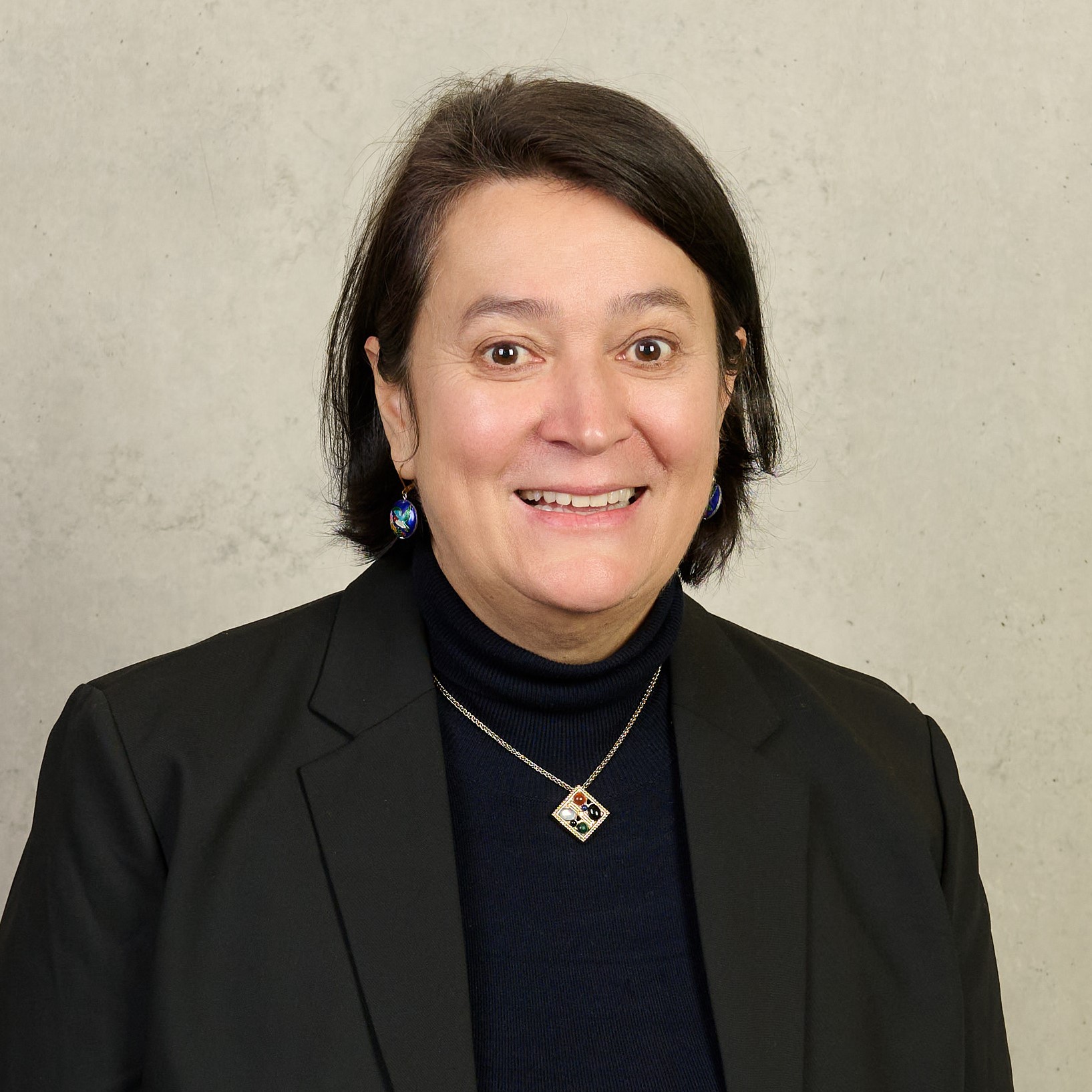 A person with shoulder-length dark hair is smiling and wearing a black blazer over a black turtleneck. They have colorful earrings and a pendant necklace. The background is a light gray textured wall.