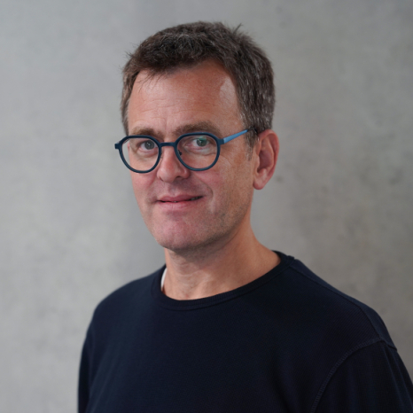 A person with short brown hair and glasses, wearing a dark shirt, stands against a plain gray background.