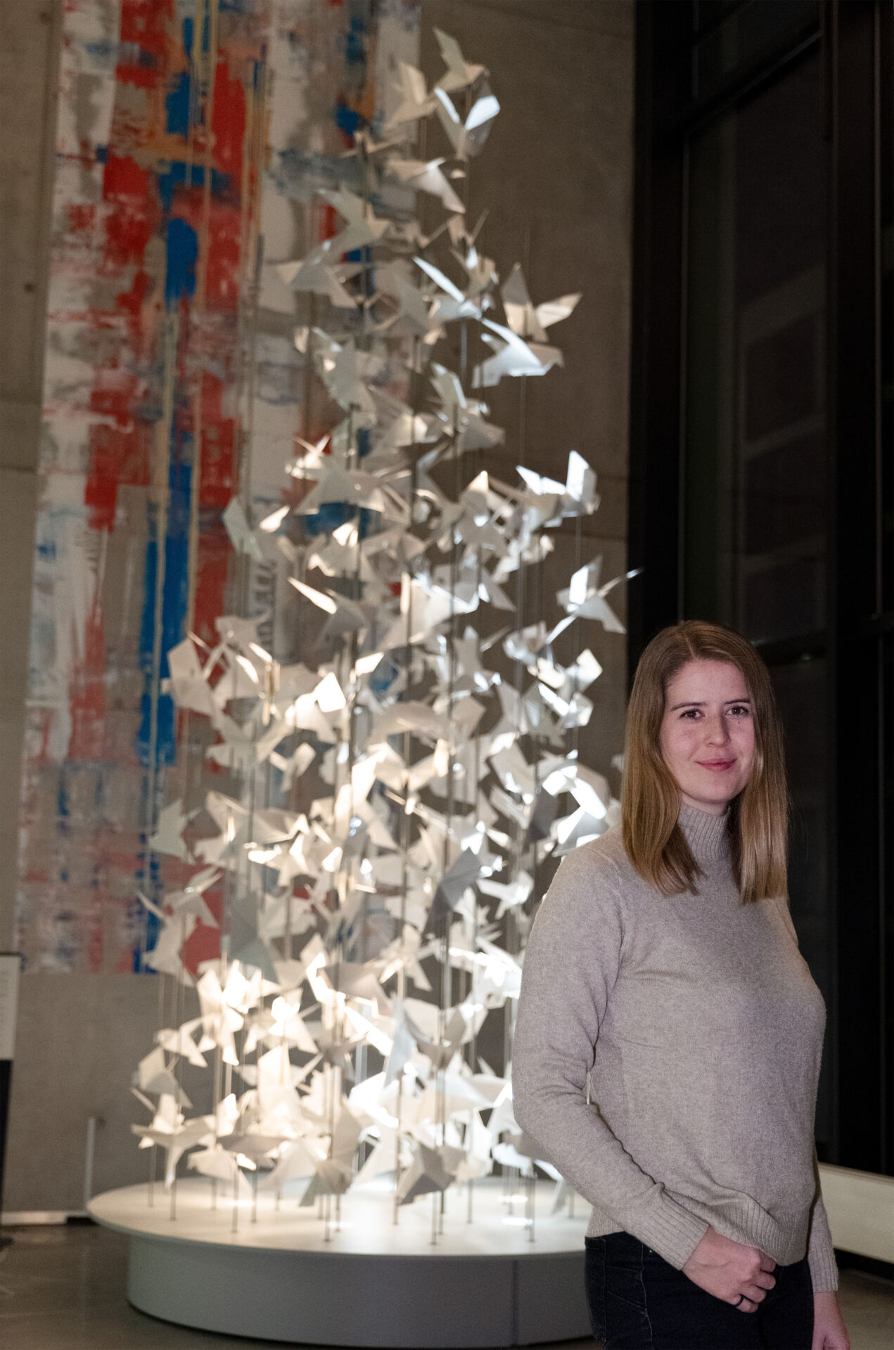 Eine Frau in einem grauen Rollkragenpullover steht vor einer modernen Skulptur mit zahlreichen weißen Papierkranichen. Hinter ihr hängt an einer hohen Wand ein großes abstraktes Gemälde in den Farben Rot, Blau und Weiß.