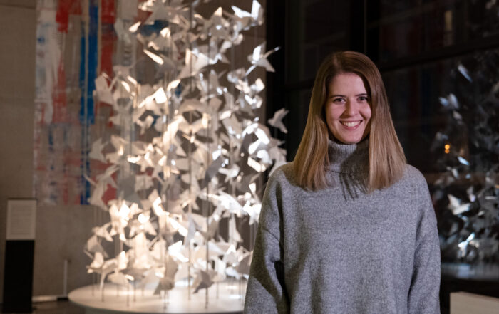 Eine Person mit langen Haaren trägt einen grauen Pullover und lächelt, während sie in einer Galerie vor einer komplizierten Kunstinstallation mit Papierkranichen steht.