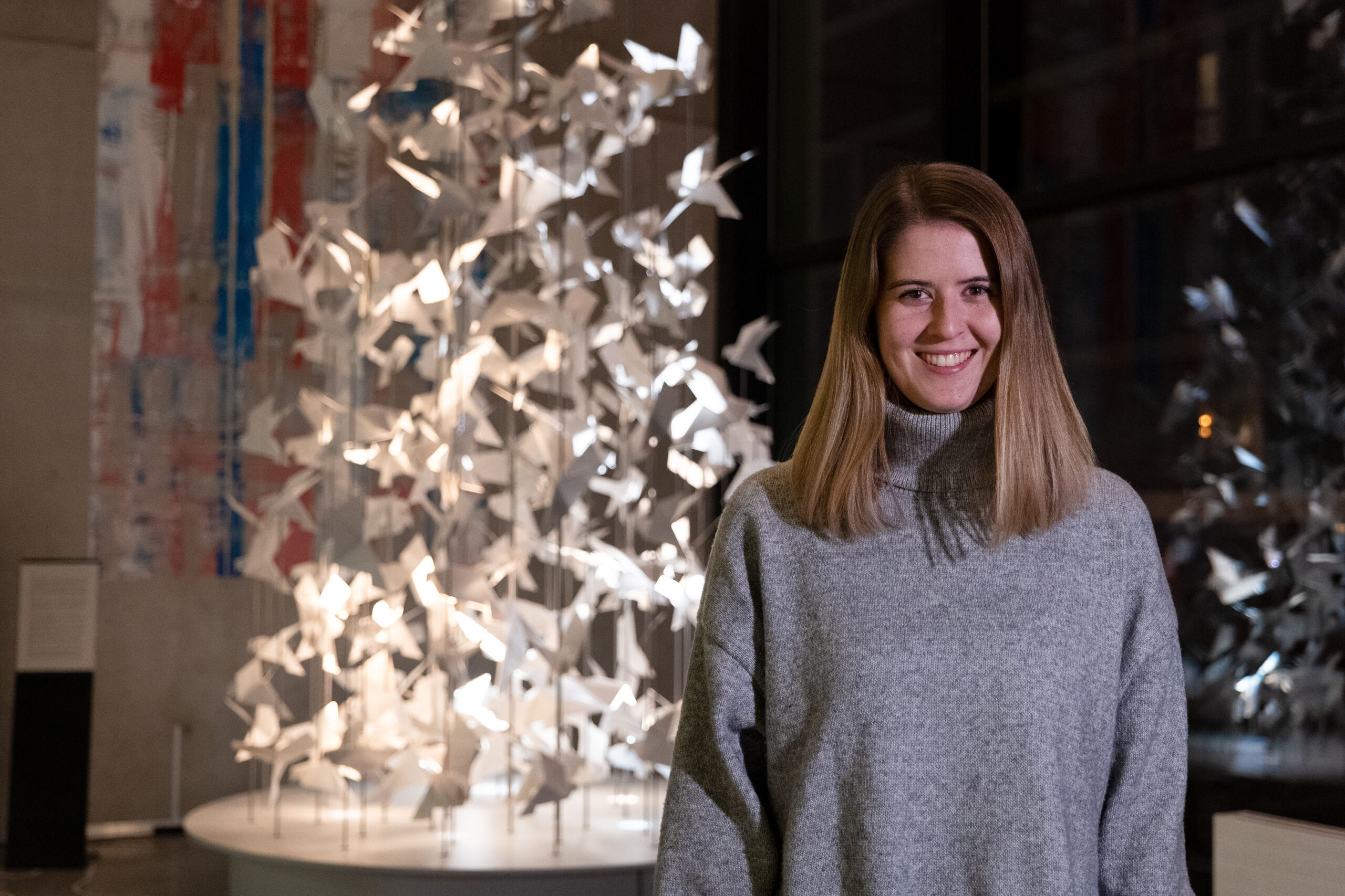 Eine Person mit langen Haaren trägt einen grauen Pullover und lächelt, während sie in einer Galerie vor einer komplizierten Kunstinstallation mit Papierkranichen steht.