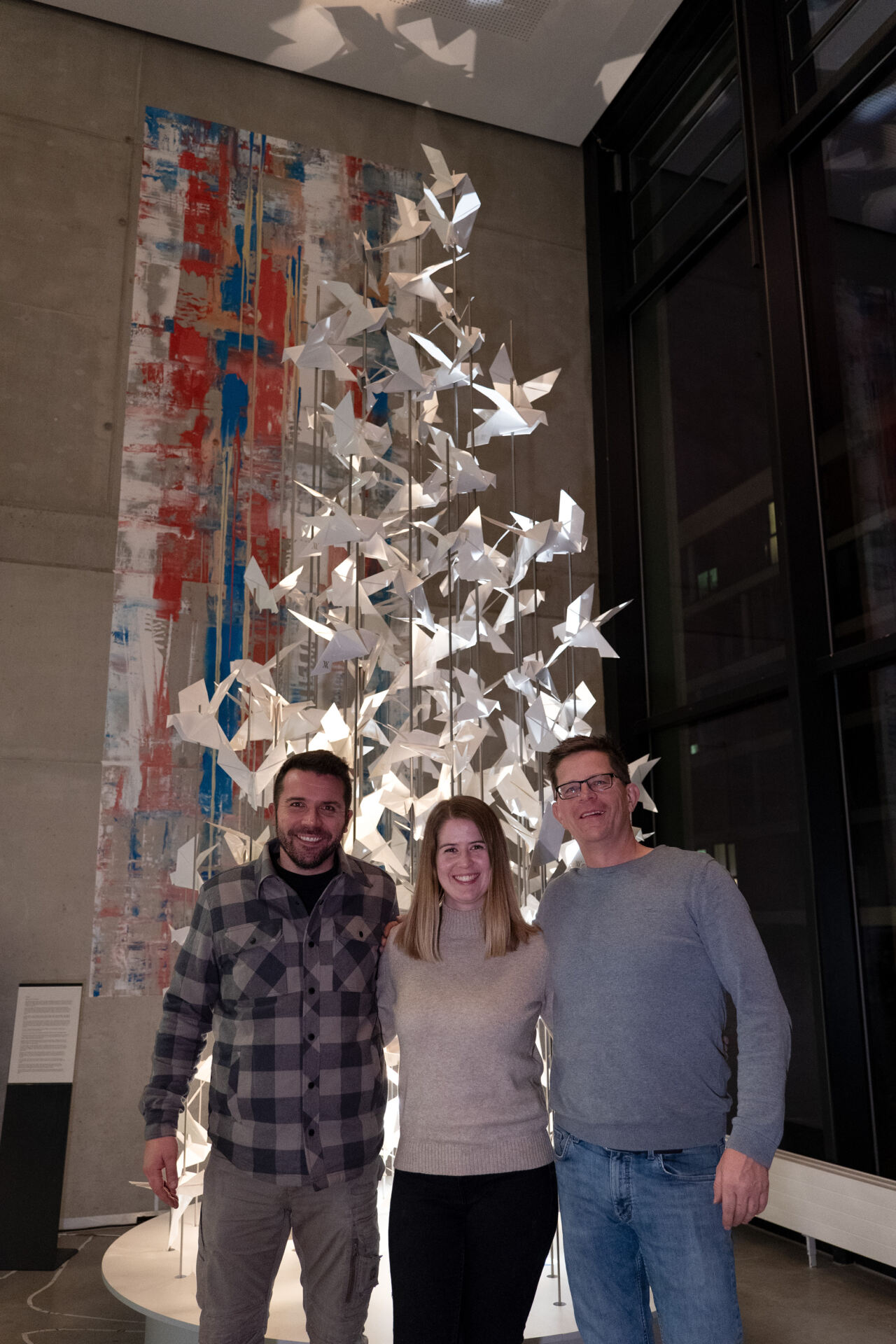 Drei Personen stehen vor einer großen, abstrakten Skulptur, die einem Baum aus Papierkranichen ähnelt. Im Hintergrund ist ein vertikales, farbenfrohes abstraktes Gemälde auf einer Betonwand zu sehen.