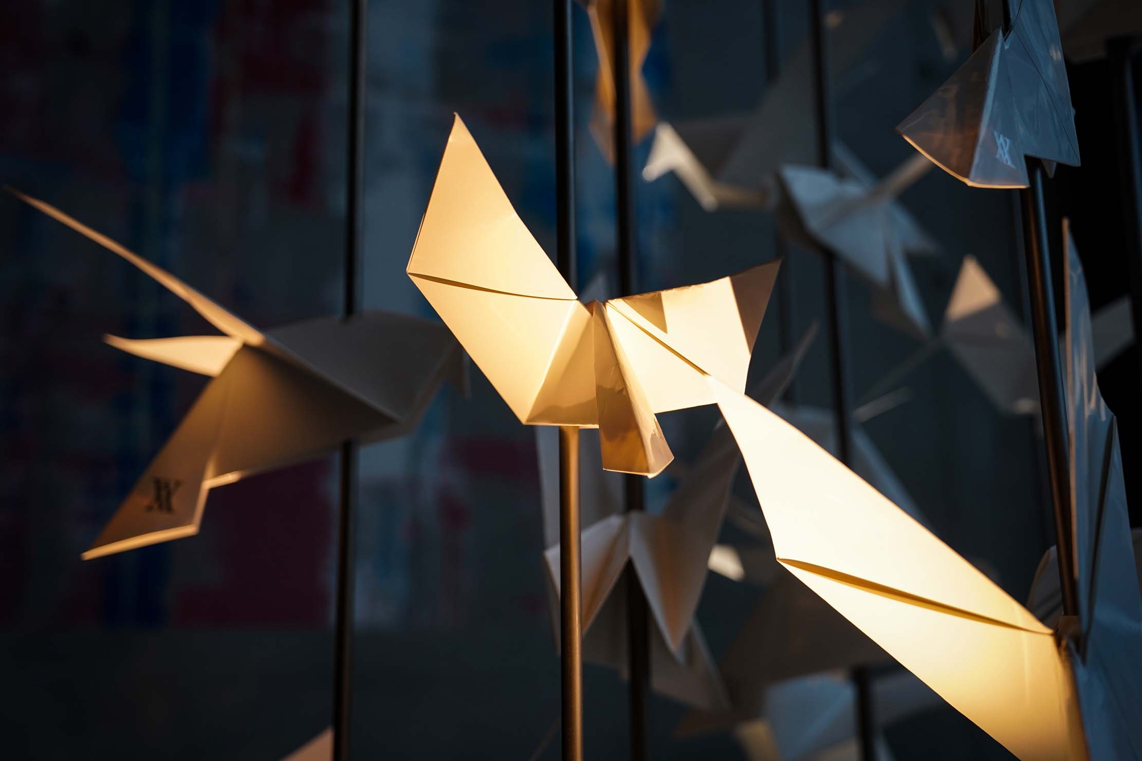 Nahaufnahme von Origami-Papiervögeln, die in der Luft schweben und von warmem Licht angestrahlt werden. Die Szene erzeugt eine ruhige Atmosphäre, wobei die zarten Falten und Schatten das kunstvolle Schauspiel vor einem unscharfen Hintergrund hervorheben.