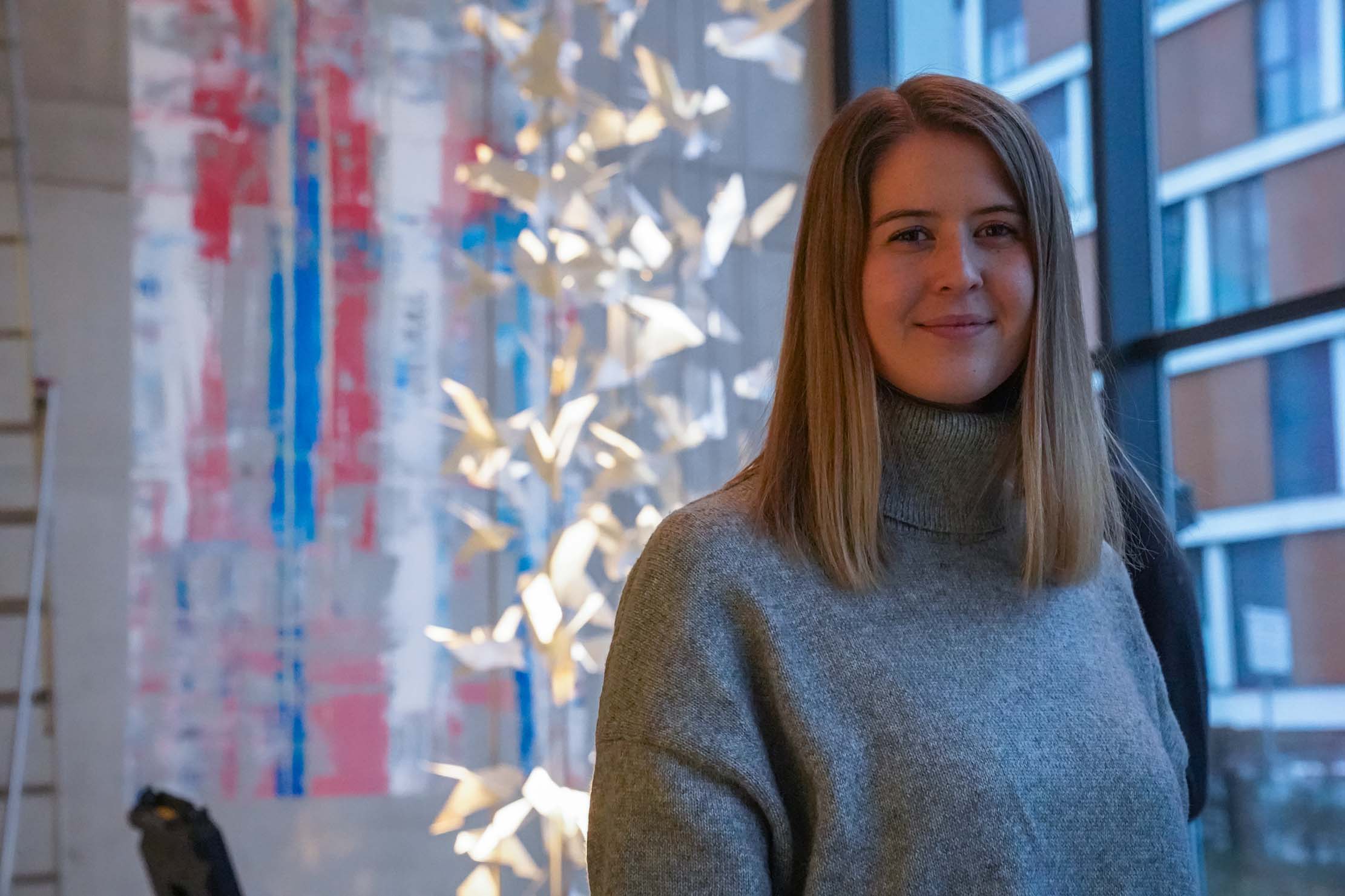 Eine Person mit langen Haaren und einem grauen Pullover steht drinnen neben einem großen Fenster. Im Hintergrund sind abstrakte Kunstwerke und ein dekorativer Baum mit weißen Blättern zu sehen.