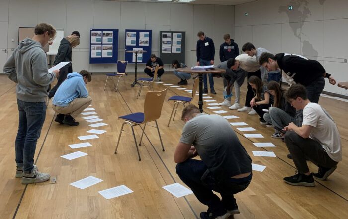 Eine Gruppe von Gründerinnen befindet sich in einem Raum mit Holzboden und beschäftigt sich mit einer Aktivität, bei der sie Papiere auf den Boden legen. Einige hocken, andere stehen und beobachten aufmerksam. Die großen, beleuchteten Deckenpaneele des Raums erhellen ihren gemeinsamen Startschuss.