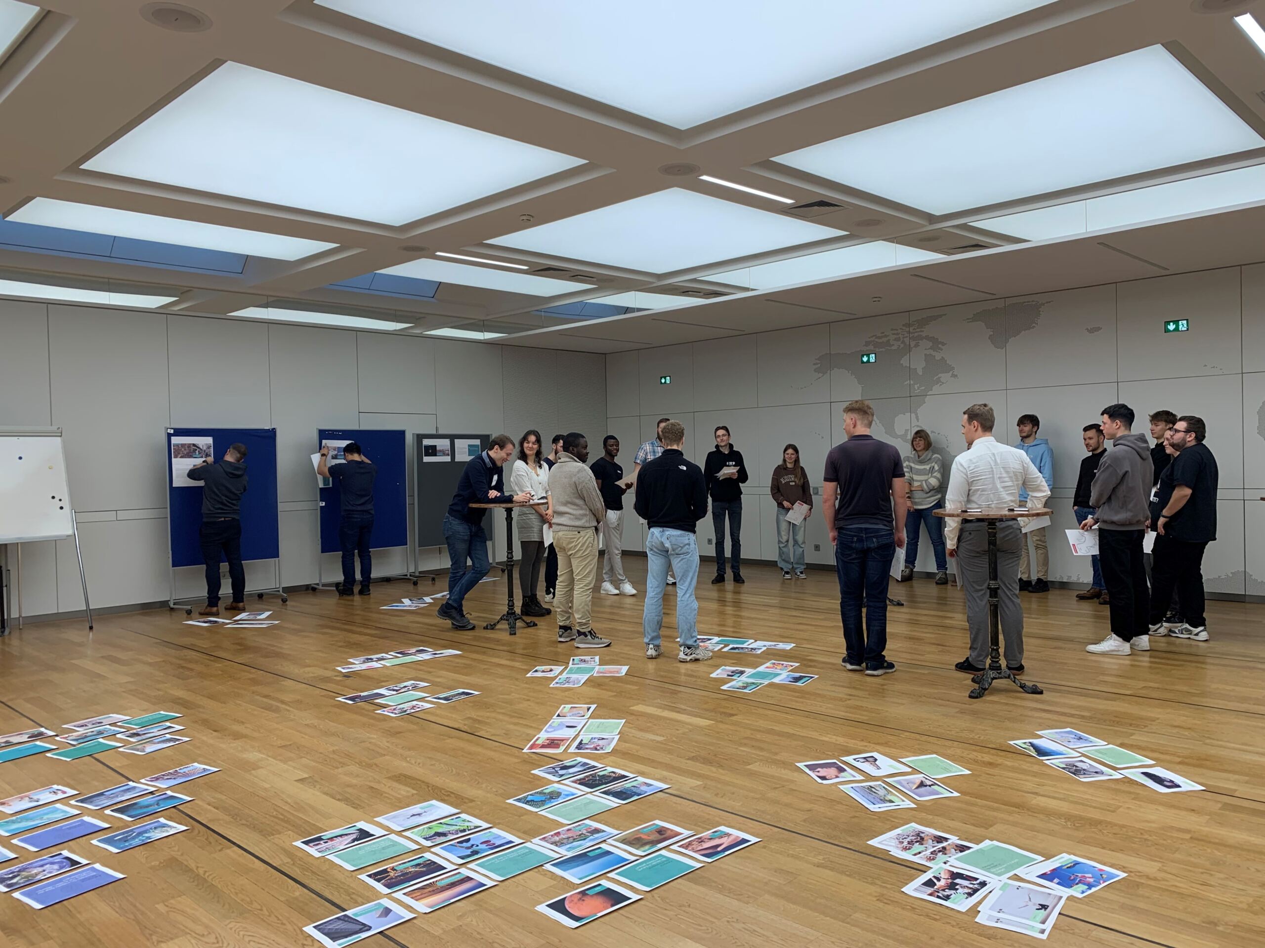 Eine Gruppe von Gründern diskutiert leidenschaftlich Ideen in einem geräumigen Raum mit Holzboden. Buntes Papiermaterial liegt verstreut herum, einiges davon ist an große Tafeln gepinnt. Oben erhellen helle quadratische Lichter die Szene und markieren den Startschuss ihrer innovativen Reise.