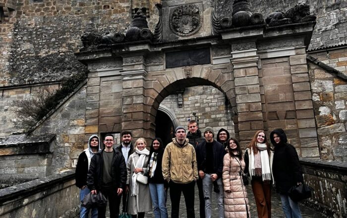 Eine Gruppe von elf Personen steht vor einer alten Steinburg in Coburg, deren gewölbter Eingang mit Wappen geschmückt ist. Der bedeckte Himmel ragt über dem gepflasterten Boden hervor, als sie sich warm angezogen für das Internationale Studierenden-Projekt versammeln. at Hochschule Coburg