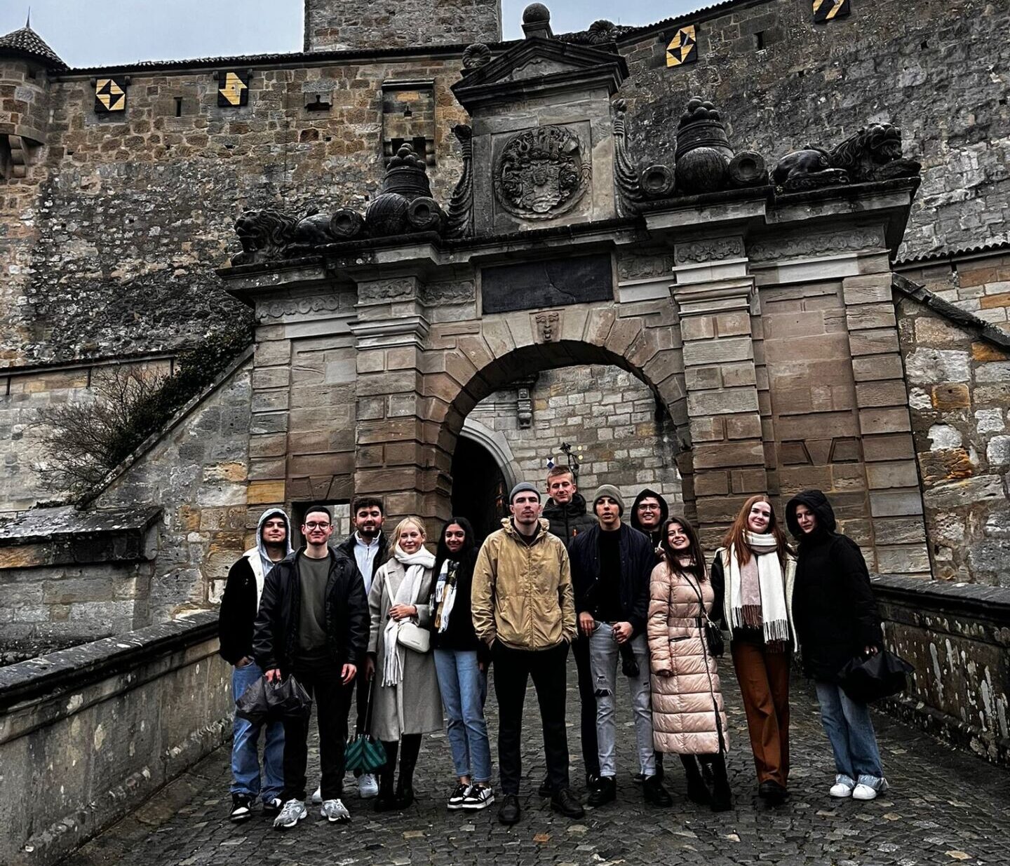 Eine Gruppe von elf Personen steht vor einer alten Steinburg in Coburg, deren gewölbter Eingang mit Wappen geschmückt ist. Der bedeckte Himmel ragt über dem gepflasterten Boden hervor, als sie sich warm angezogen für das Internationale Studierenden-Projekt versammeln. at Hochschule Coburg