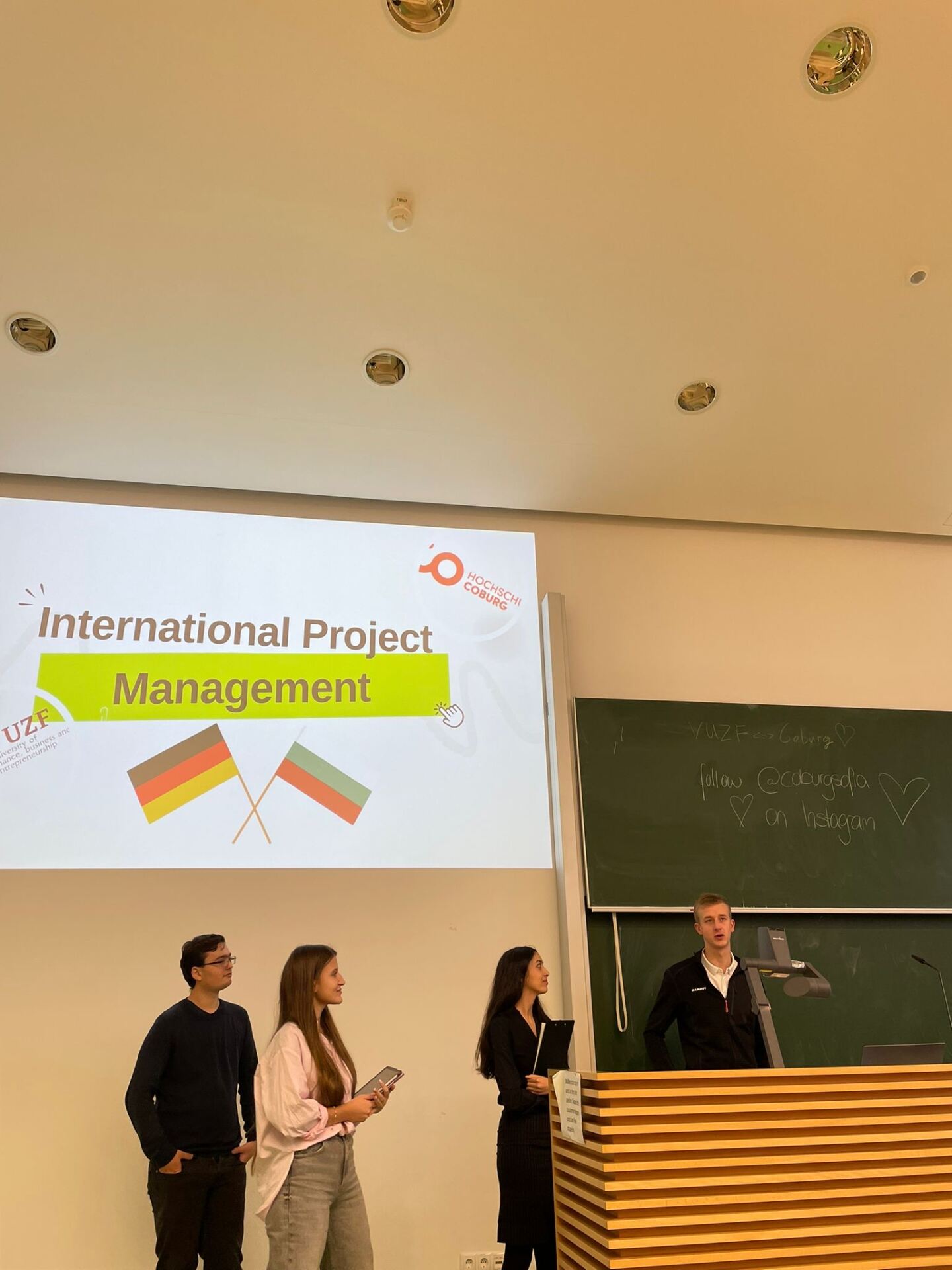 Vier Personen stehen in einem Hörsaal und halten im Rahmen eines Studierenden-Projekts einen Vortrag zum Thema „Internationales Projektmanagement“. Die deutsche und die bulgarische Flagge zieren die Folie und deuten auf eine lebendige Zusammenarbeit zwischen Berlin und Sofia hin. Im Hintergrund ist eine Tafel teilweise sichtbar. at Hochschule Coburg