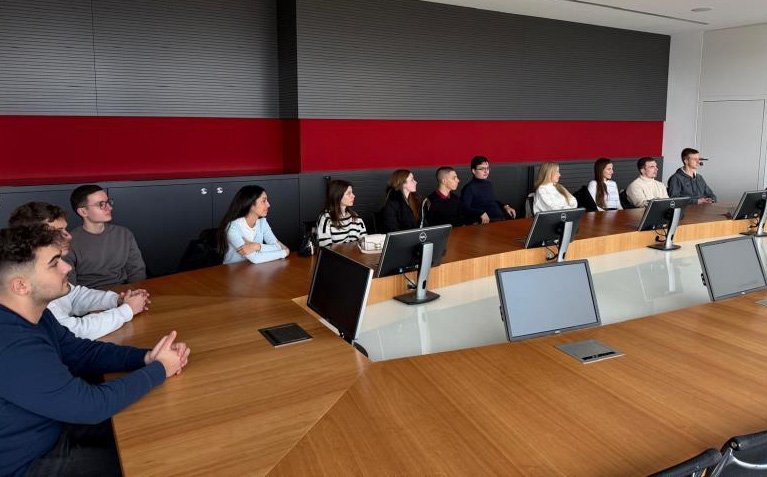 Eine Gruppe von Menschen sitzt an einem langen Konferenztisch mit Bildschirmen vor sich und schaut einem Vortrag oder einer Präsentation zum Internationalen Studierenden-Projekt zu. Der Raum ist modern gestaltet, mit roten und schwarzen Wänden, die an die Wände in Coburg erinnern. Die Teilnehmer hören aufmerksam zu. at Hochschule Coburg