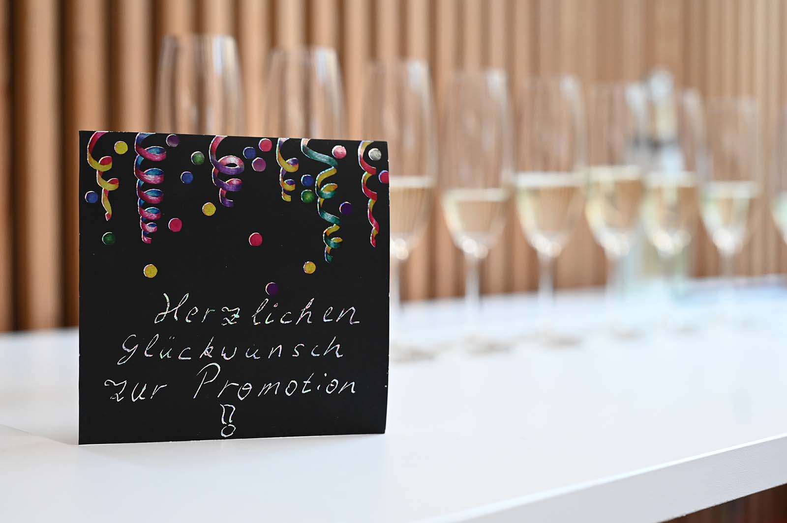 Auf einem weißen Tisch steht eine Glückwunschkarte mit „Herzlichen Glückwunsch zur Promotion“ und bunten Dekorationen, die einem automatisch gespeicherten Entwurf gleicht. Im Hintergrund sorgen mehrere mit Champagner gefüllte Gläser für festliche Stimmung. at Hochschule Coburg