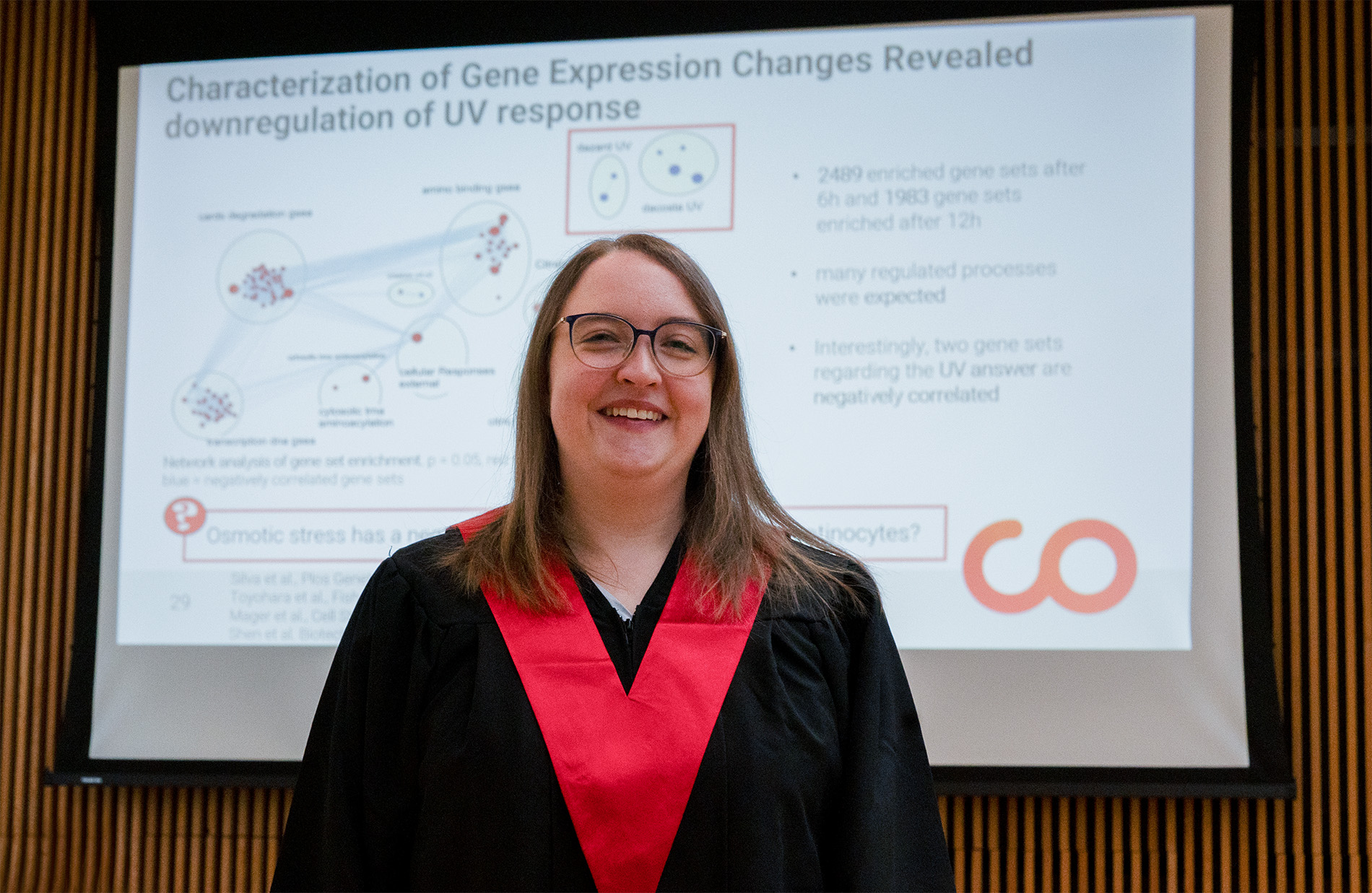 In akademischer Kleidung lächelt eine Person vor einer Präsentationsfolie mit dem Titel "... die Genexpression und UV-Reaktion untersucht. Die Folie enthält Diagramme und Text, die angereicherte Gensätze im Zusammenhang mit osmotischem Stress detailliert beschreiben. at Hochschule Coburg