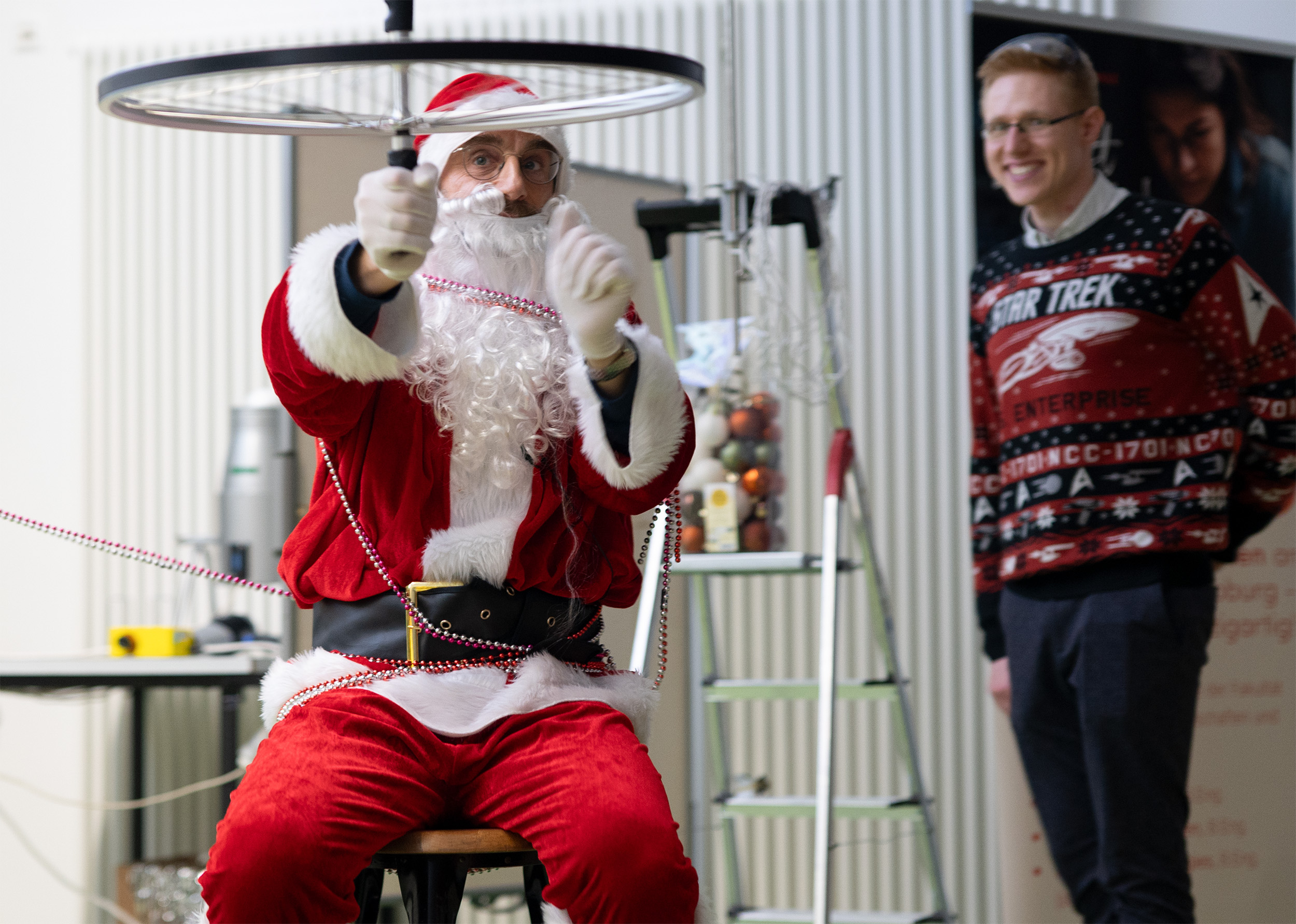 Während einer Weihnachtsvorlesung balanciert jemand, der als Weihnachtsmann verkleidet ist, eindrucksvoll ein sich drehendes Fahrradrad auf einem Stock. In der Nähe lächelt eine andere Person in einem festlichen Pullover und verkörpert die Weihnachtsstimmung. Die Szene ist voller Fröhlichkeit und wahrscheinlich Teil einer fröhlichen Weihnachtsveranstaltung. at Hochschule Coburg