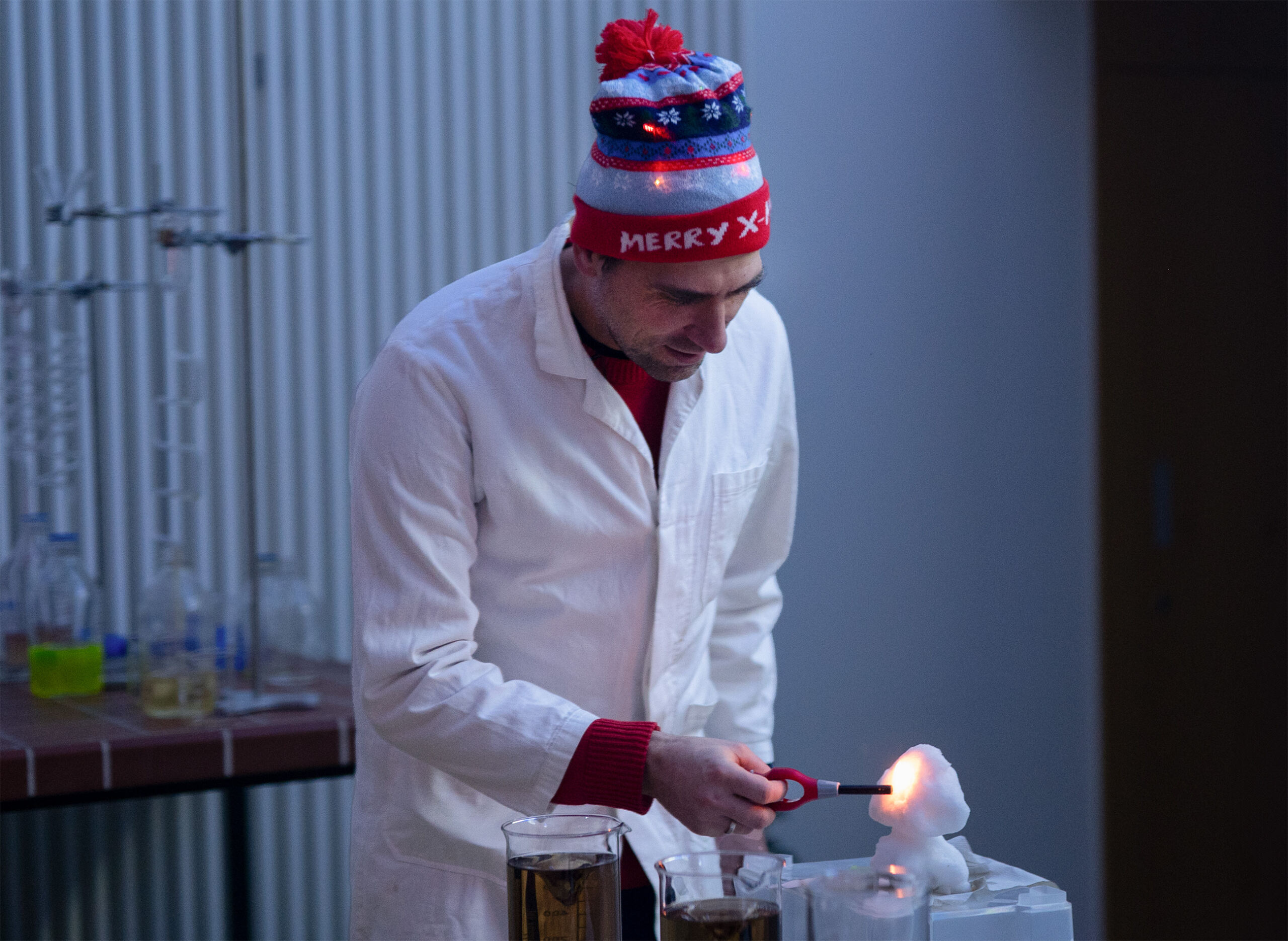 Eine Person mit „Merry Xmas“-Mütze und Laborkittel führt ein Weihnachtsvorlesungsexperiment durch, bei dem sie im Labor ein Feuerzeug an einem Schneemann aus Schaum oder Schnee anzündet. Becher mit Flüssigkeit stehen verstreut auf einem Tisch in der Nähe und tragen zur festlichen Wissenschaftsszene bei. at Hochschule Coburg