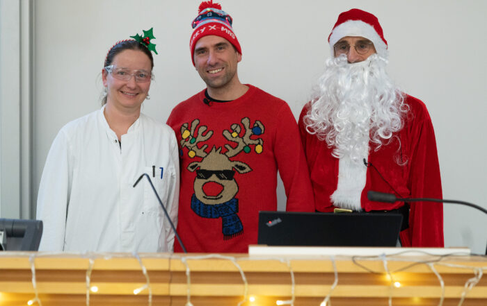 Drei Personen in festlicher Weihnachtskleidung versammeln sich hinter einem mit Lichterketten geschmückten Holzpodest. Einer trägt einen Laborkittel und ein Rentiergeweih, ein anderer einen Rentierpullover und eine Wintermütze, und Weihnachts-Stefan vervollständigt die Szene in seinem Weihnachtsmannkostüm und seiner Mütze. at Hochschule Coburg