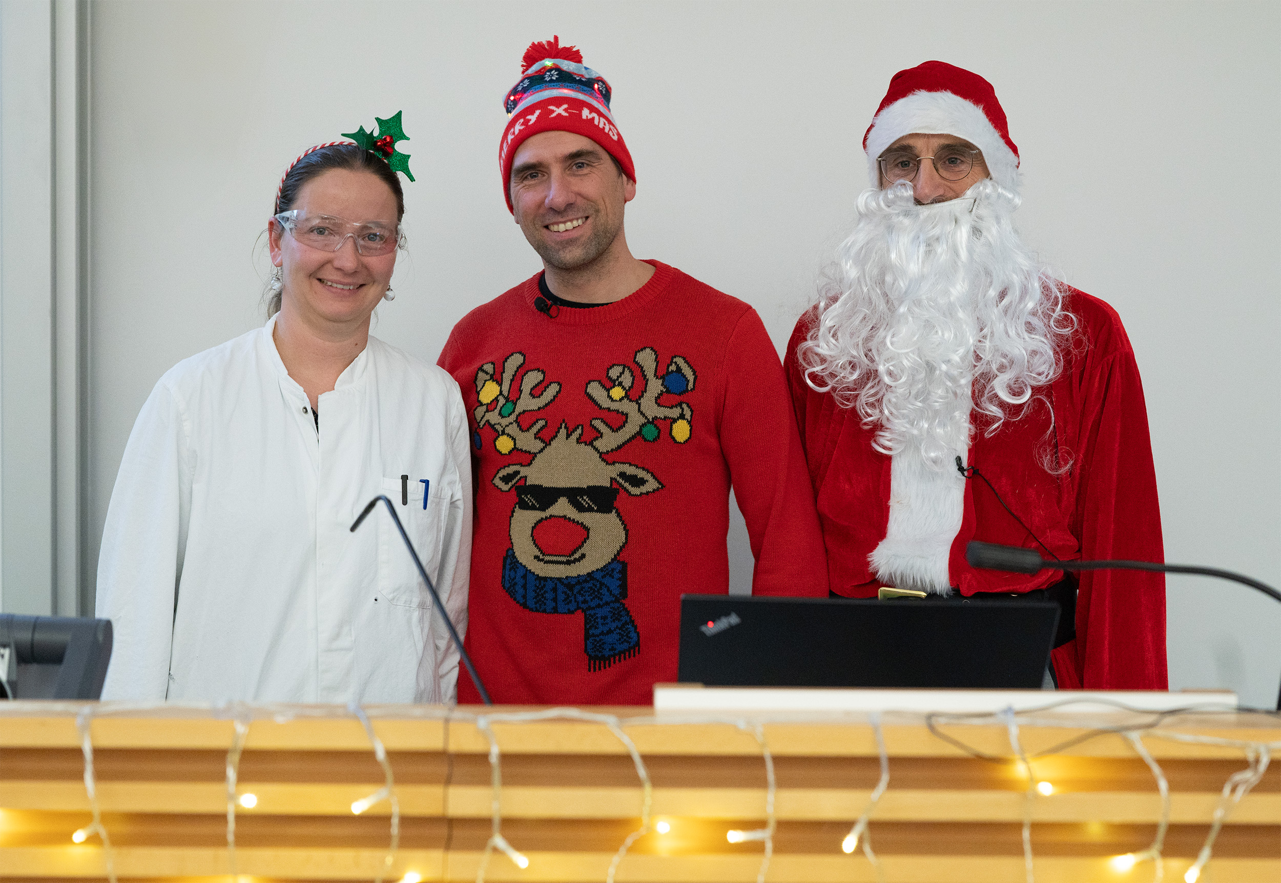 Drei Personen in festlicher Weihnachtskleidung versammeln sich hinter einem mit Lichterketten geschmückten Holzpodest. Einer trägt einen Laborkittel und ein Rentiergeweih, ein anderer einen Rentierpullover und eine Wintermütze, und Weihnachts-Stefan vervollständigt die Szene in seinem Weihnachtsmannkostüm und seiner Mütze. at Hochschule Coburg