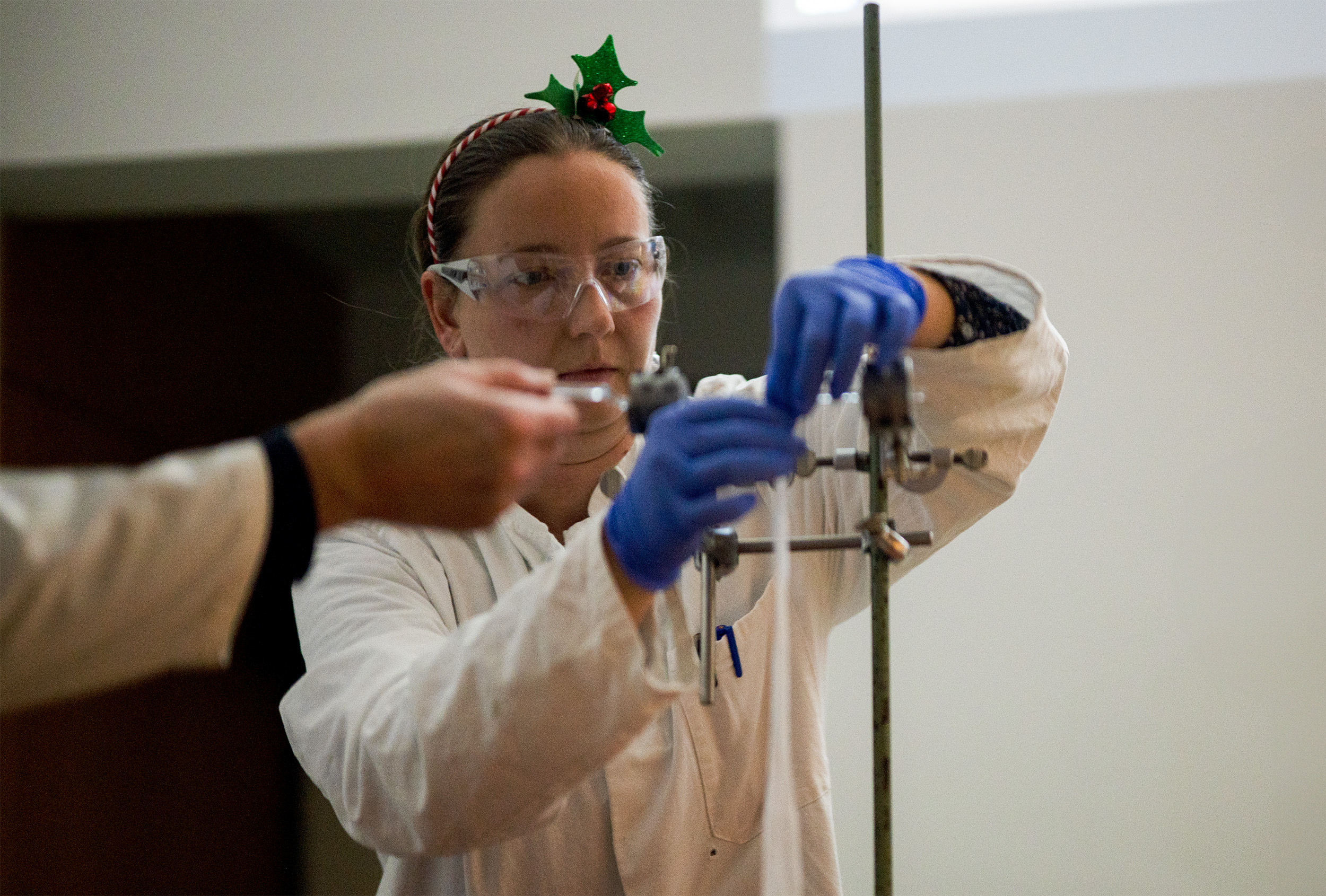 Eine Person mit einem festlichen Stirnband, das an den Weihnachtsmann erinnert, und einer Schutzbrille arbeitet in einem Labor und stellt mit blauen Handschuhen Geräte ein. Eine weitere Person, die möglicherweise an der Weihnachtsvorlesung beteiligt ist, ist im Vordergrund teilweise sichtbar und trägt ebenfalls einen Laborkittel. at Hochschule Coburg