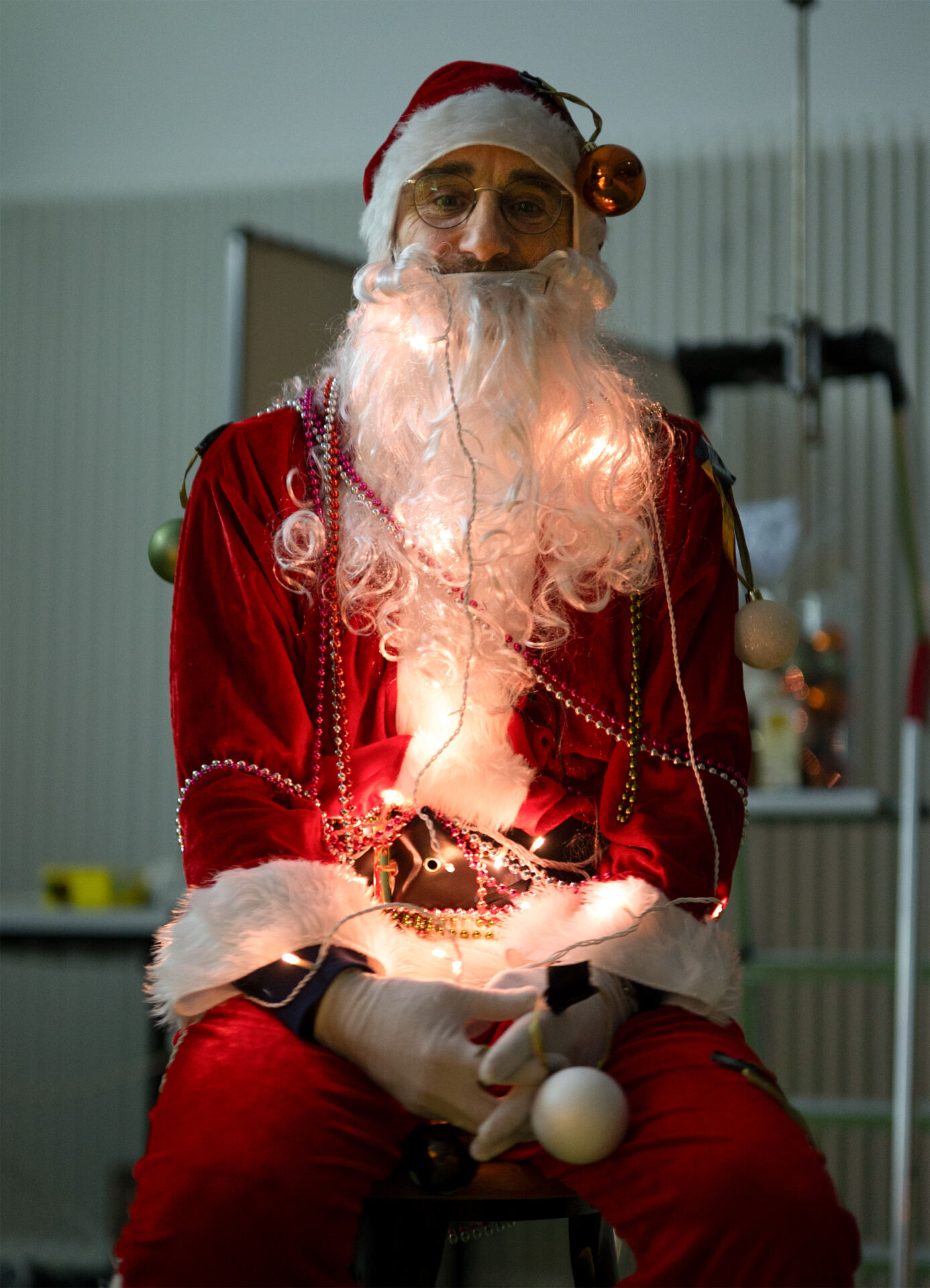 Eine als Weihnachtsmann verkleidete Person sitzt mit den Händen im Schoß und trägt ein rotes Kostüm mit weißem Pelzbesatz. Das Outfit funkelt mit Lichtern und Kugeln und ergänzt den langen weißen Bart. In der festlichen Stimmung von X-Mas-Stefan deutet der verschwommene Hintergrund auf Feiertagsstimmung hin. at Hochschule Coburg