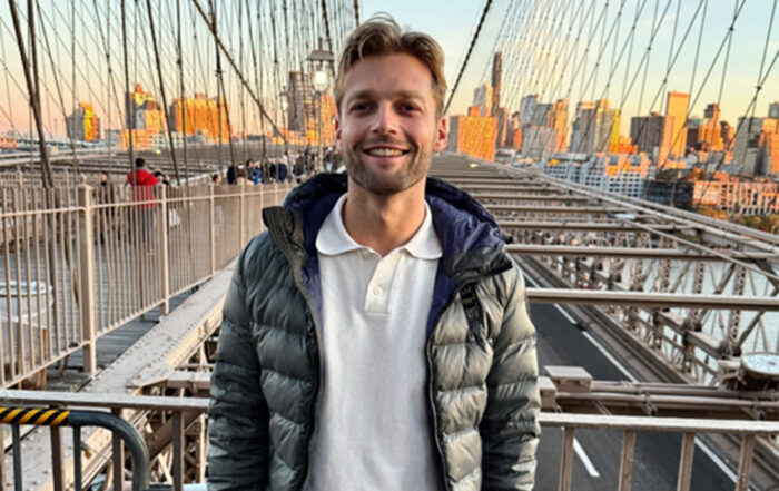 Ein BWL-Alumnus in grauer Pufferjacke und weißem Hemd steht lächelnd auf einer Brücke in New York, vor der sich die Wolkenkratzer der Stadt im Sonnenuntergang abzeichnen. Die Kabel der Brücke bilden ein markantes geometrisches Muster um sie herum. at Hochschule Coburg