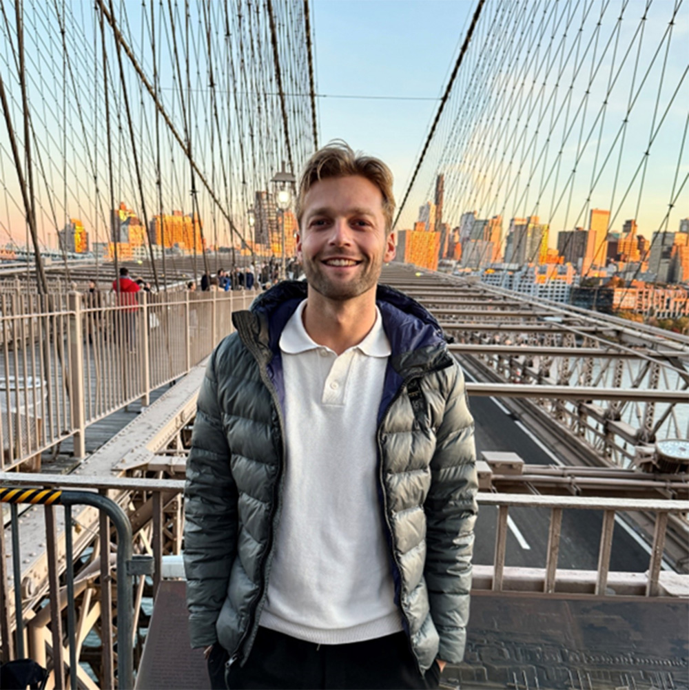 Ein BWL-Alumnus in grauer Pufferjacke und weißem Hemd steht lächelnd auf einer Brücke in New York, vor der sich die Wolkenkratzer der Stadt im Sonnenuntergang abzeichnen. Die Kabel der Brücke bilden ein markantes geometrisches Muster um sie herum. at Hochschule Coburg