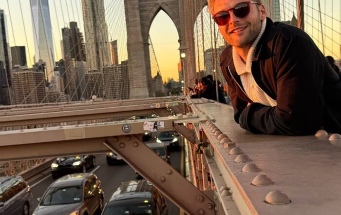 Ein BWL-Alumnus mit Sonnenbrille lehnt lächelnd am Geländer der Brooklyn Bridge. Die Sonne geht unter und taucht New York in einen warmen Glanz. Unterhalb der Straße rollen Autos vorbei, die Skyline der Stadt bildet eine malerische Kulisse, die an einen Abend in Übersee erinnert. at Hochschule Coburg