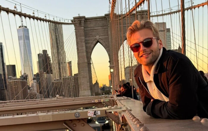 Ein BWL-Alumnus mit Sonnenbrille lehnt lächelnd am Geländer der Brooklyn Bridge. Die Sonne geht unter und taucht New York in einen warmen Glanz. Unterhalb der Straße rollen Autos vorbei, die Skyline der Stadt bildet eine malerische Kulisse, die an einen Abend in Übersee erinnert. at Hochschule Coburg