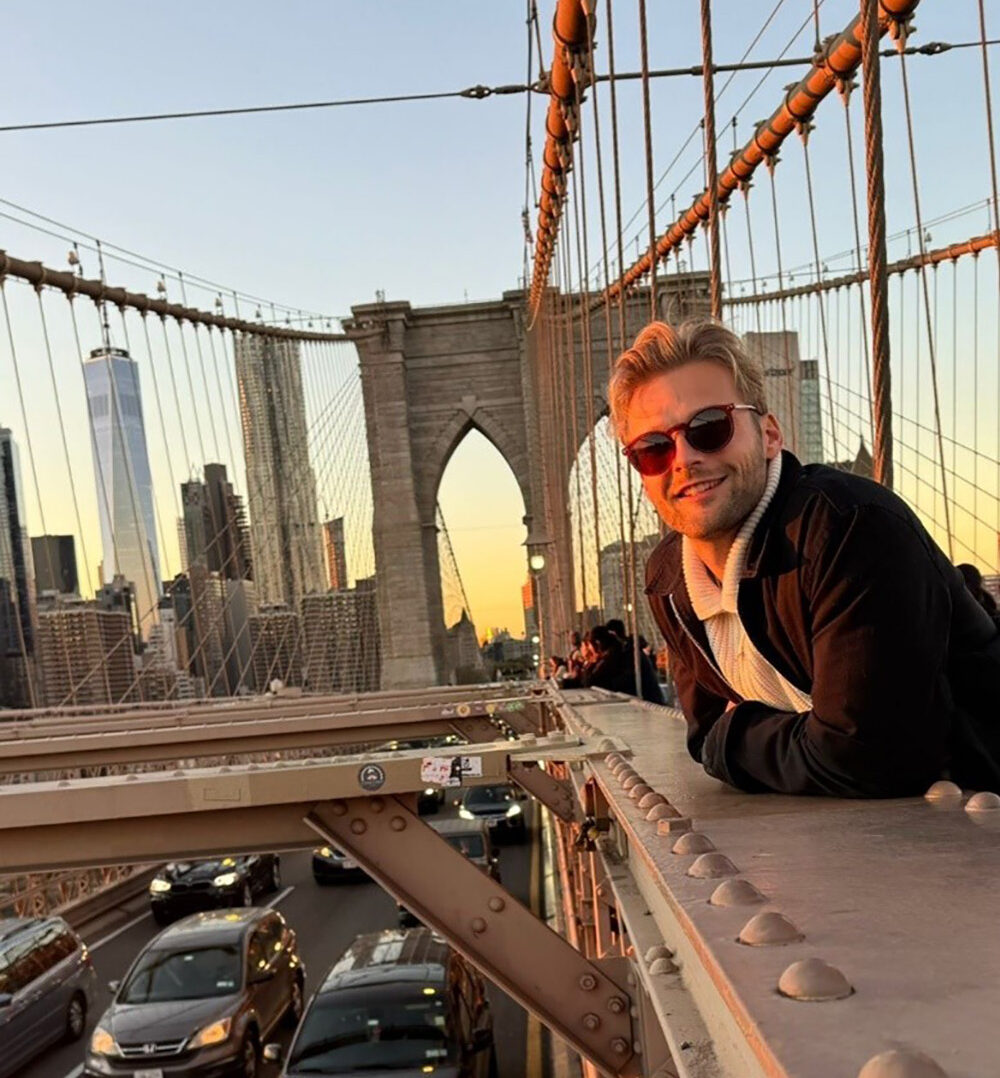 Ein BWL-Alumnus mit Sonnenbrille lehnt lächelnd am Geländer der Brooklyn Bridge. Die Sonne geht unter und taucht New York in einen warmen Glanz. Unterhalb der Straße rollen Autos vorbei, die Skyline der Stadt bildet eine malerische Kulisse, die an einen Abend in Übersee erinnert. at Hochschule Coburg