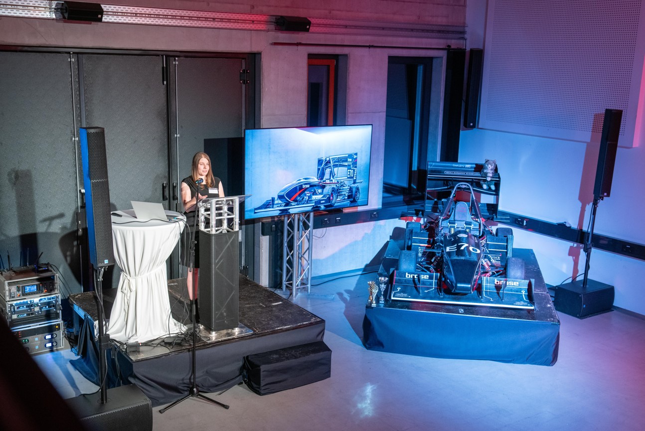 Eine Frau präsentiert auf einem Podium mit einem großen Monitor, auf dem die Blaupause eines Rennwagens angezeigt wird. Neben ihr wird auf einer Plattform ein echtes Rennauto ausgestellt. Die Kulisse ist ein Innenraum mit technischer Ausstattung und Beleuchtung.