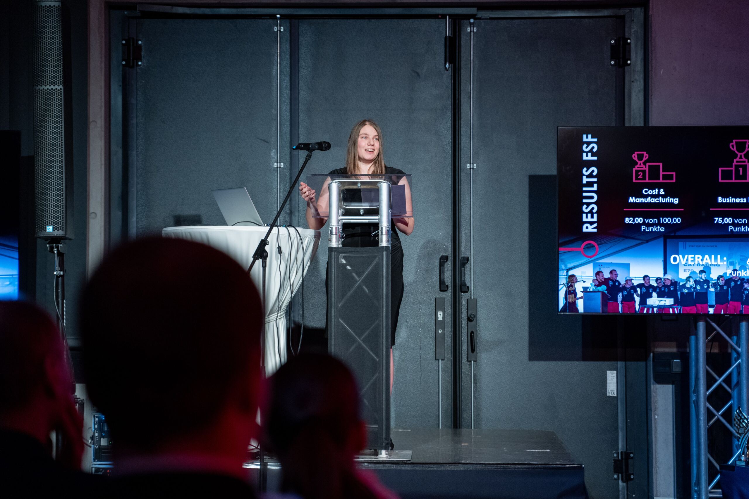 Eine Frau steht mit einem Mikrofon an einem Podium und hält eine Präsentation. Hinter ihr ist ein Bildschirm zu sehen, auf dem Ergebnisse und Kategorien wie „Kosten & Herstellung“ und „Geschäftswert“ angezeigt werden. Im Vordergrund sitzen die Zuhörer.