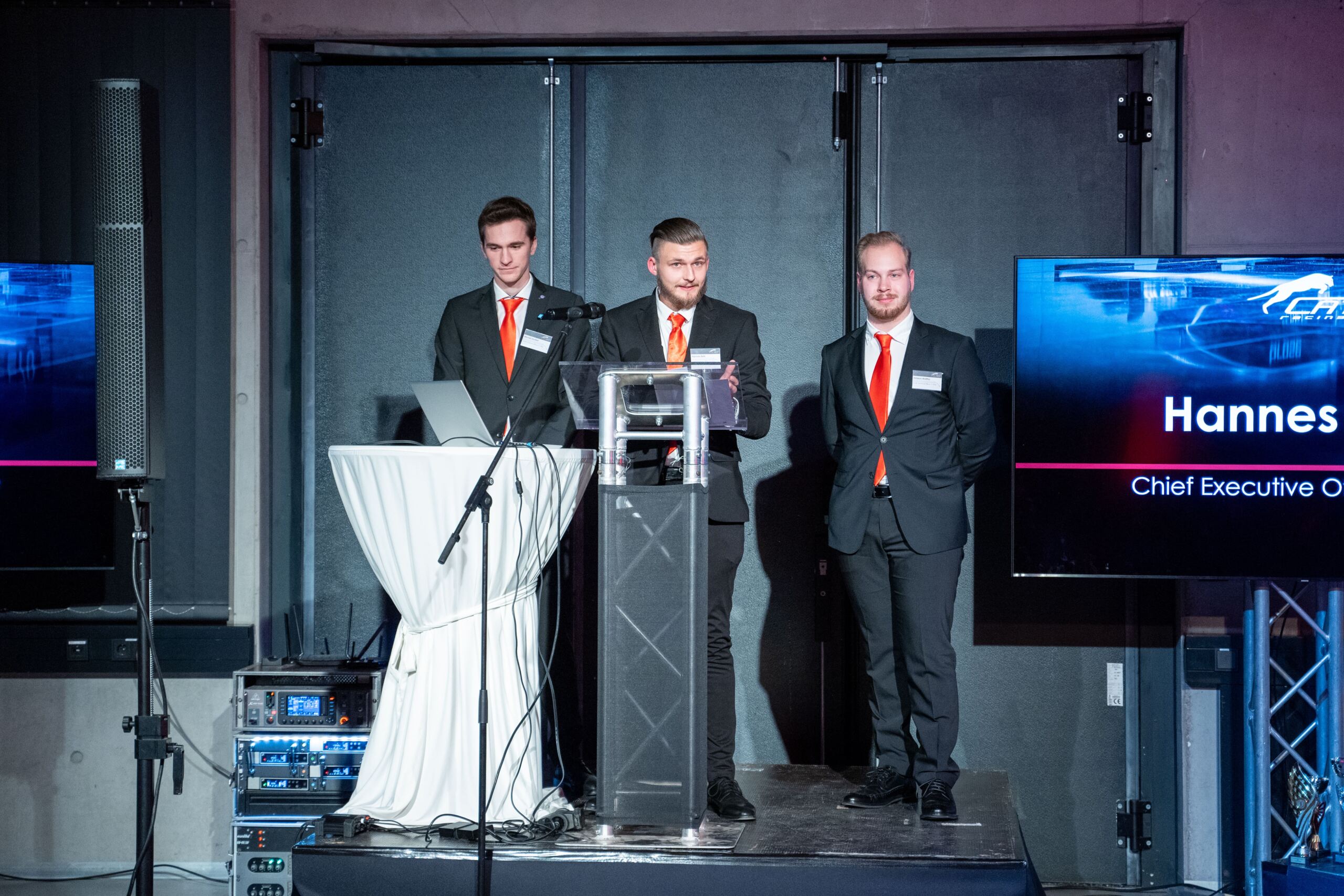 Drei Männer in Anzügen und mit roten Krawatten stehen hinter einem Podium auf einer Bühne. Einer liest von einem Laptop ab, ein anderer spricht in ein Mikrofon und der dritte steht mit gefalteten Händen da. Neben ihnen wird auf einem Bildschirm Text angezeigt.