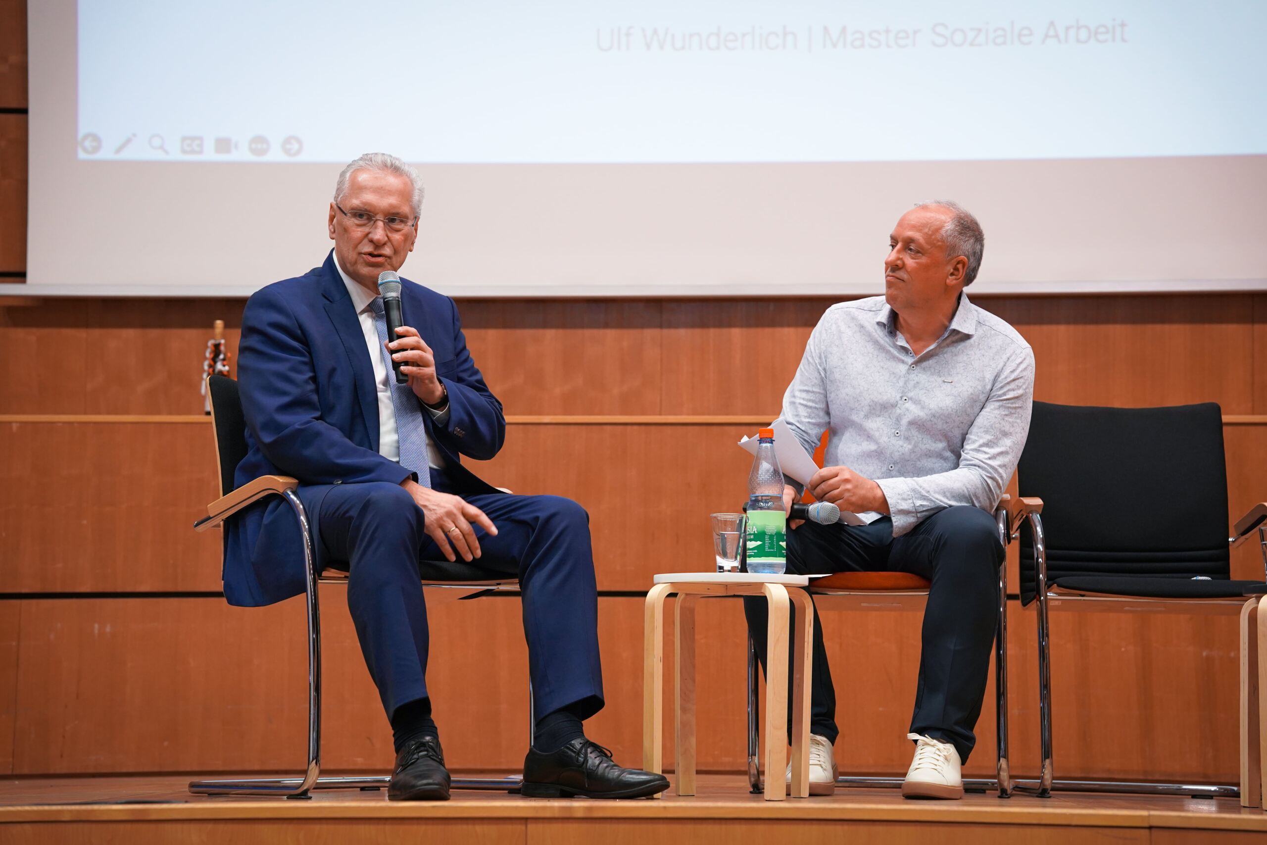 Zwei Männer sitzen auf einer Bühne und unterhalten sich. Der Mann links hält ein Mikrofon, während der Mann rechts einen Stift und einen Notizblock hält. Sie sitzen vor einem hölzernen Hintergrund, über ihnen ist eine Projektionsleinwand.