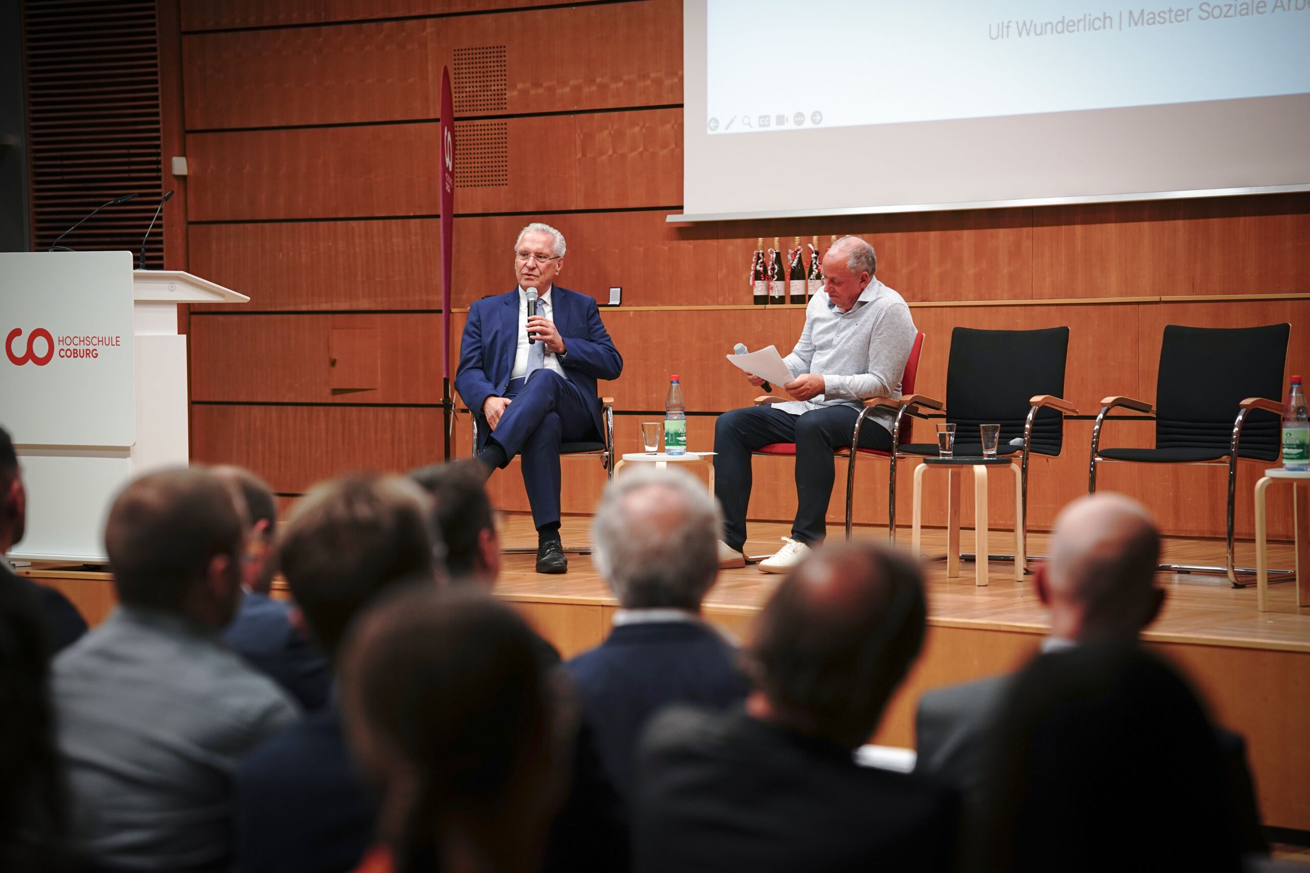 Zwei Männer sitzen auf einer Bühne und nehmen an einer Diskussion teil. Einer hält ein Mikrofon, während der andere Dokumente prüft. Ein Publikum schaut zu. Die Veranstaltung findet an der Hochschule Coburg statt. Links ist ein Rednerpult zu sehen.