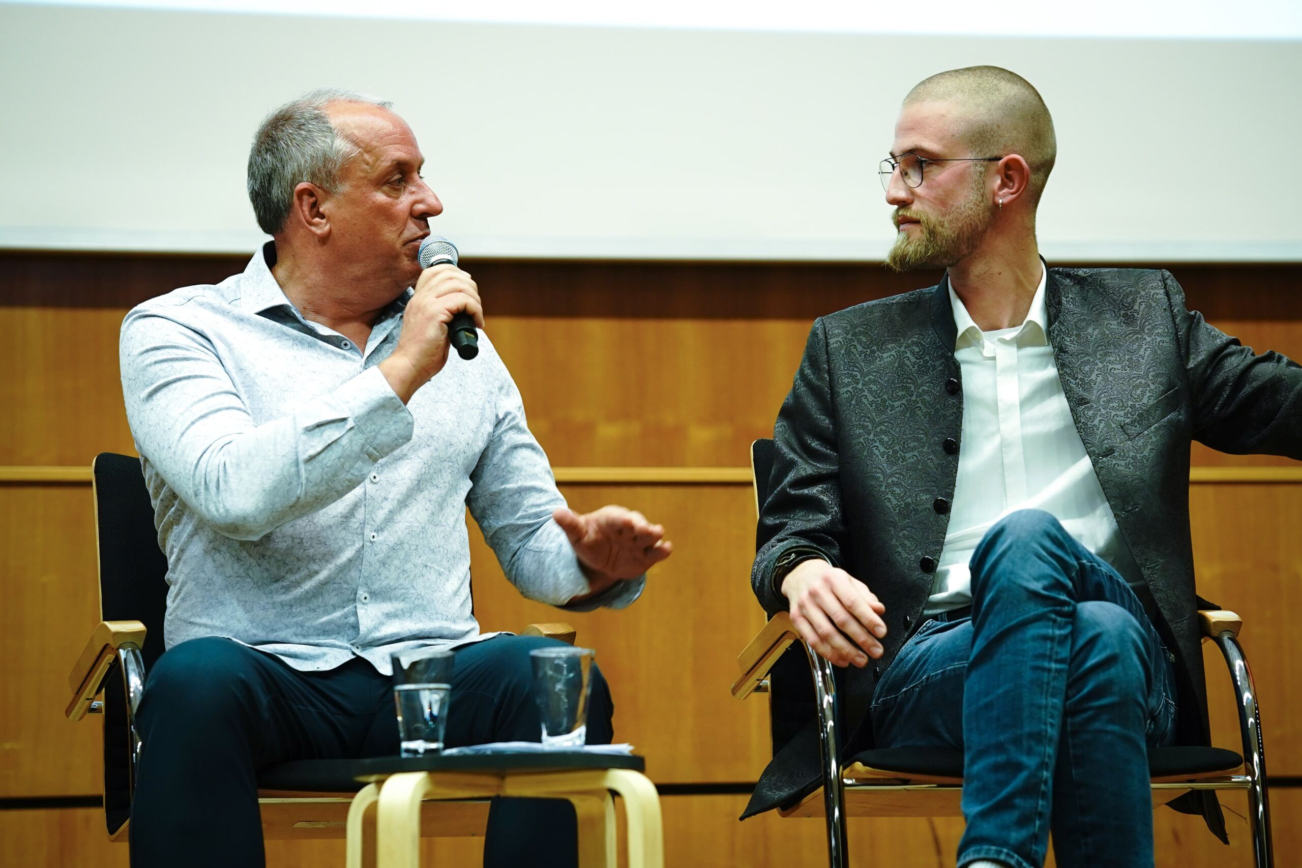 Zwei Männer sitzen auf einer Bühne und unterhalten sich. Der Mann links hält ein Mikrofon und trägt ein helles Hemd. Der Mann rechts, mit Brille und dunkler Jacke, hört aufmerksam zu. Eine Holzwand und eine Leinwand im Hintergrund sind zu sehen.