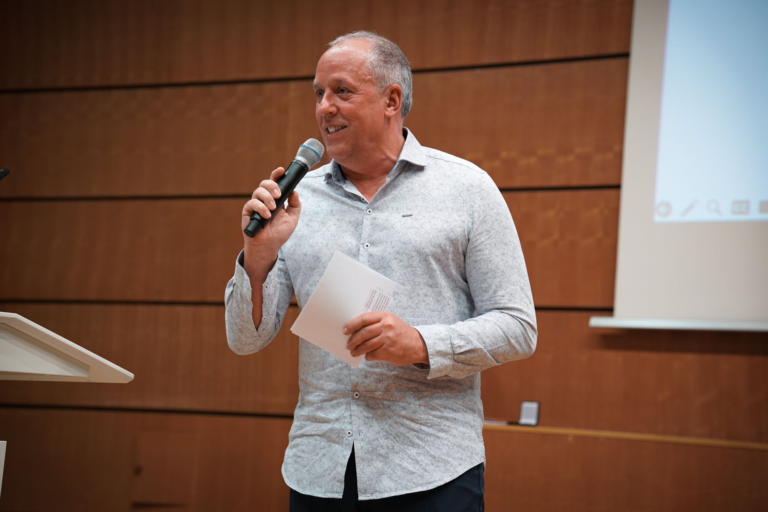 Ein Mann in einem hellgrauen Hemd spricht in ein Mikrofon, während er Papiere hält und an einem Podium vor einer Holzwand steht.