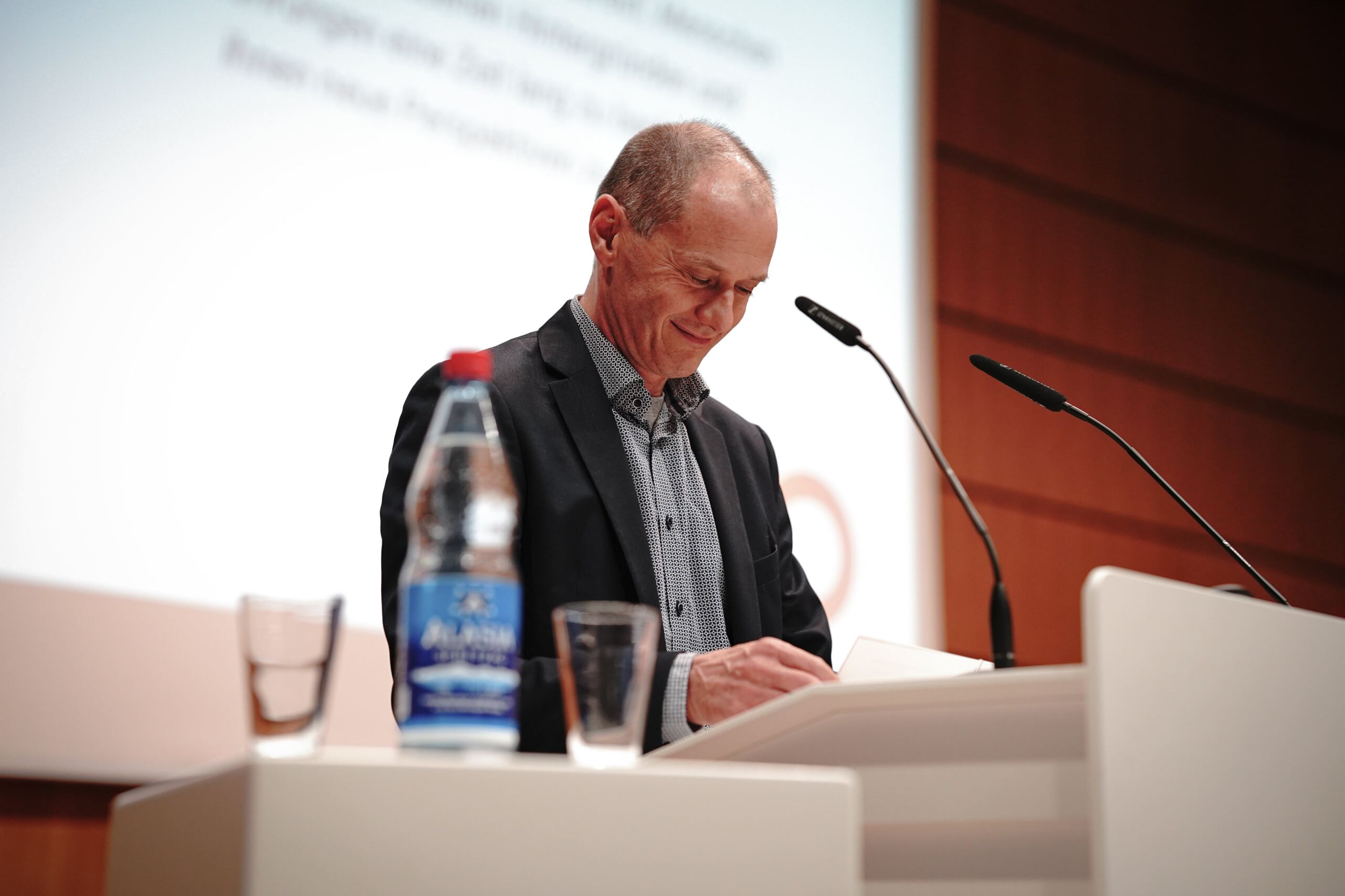 Ein Mann im Anzug steht an einem Podium mit einem Mikrofon und blickt auf Unterlagen. Auf dem Podium stehen eine Flasche Wasser und ein Glas. Im Hintergrund ist ein teilweise sichtbarer Präsentationsbildschirm zu sehen.