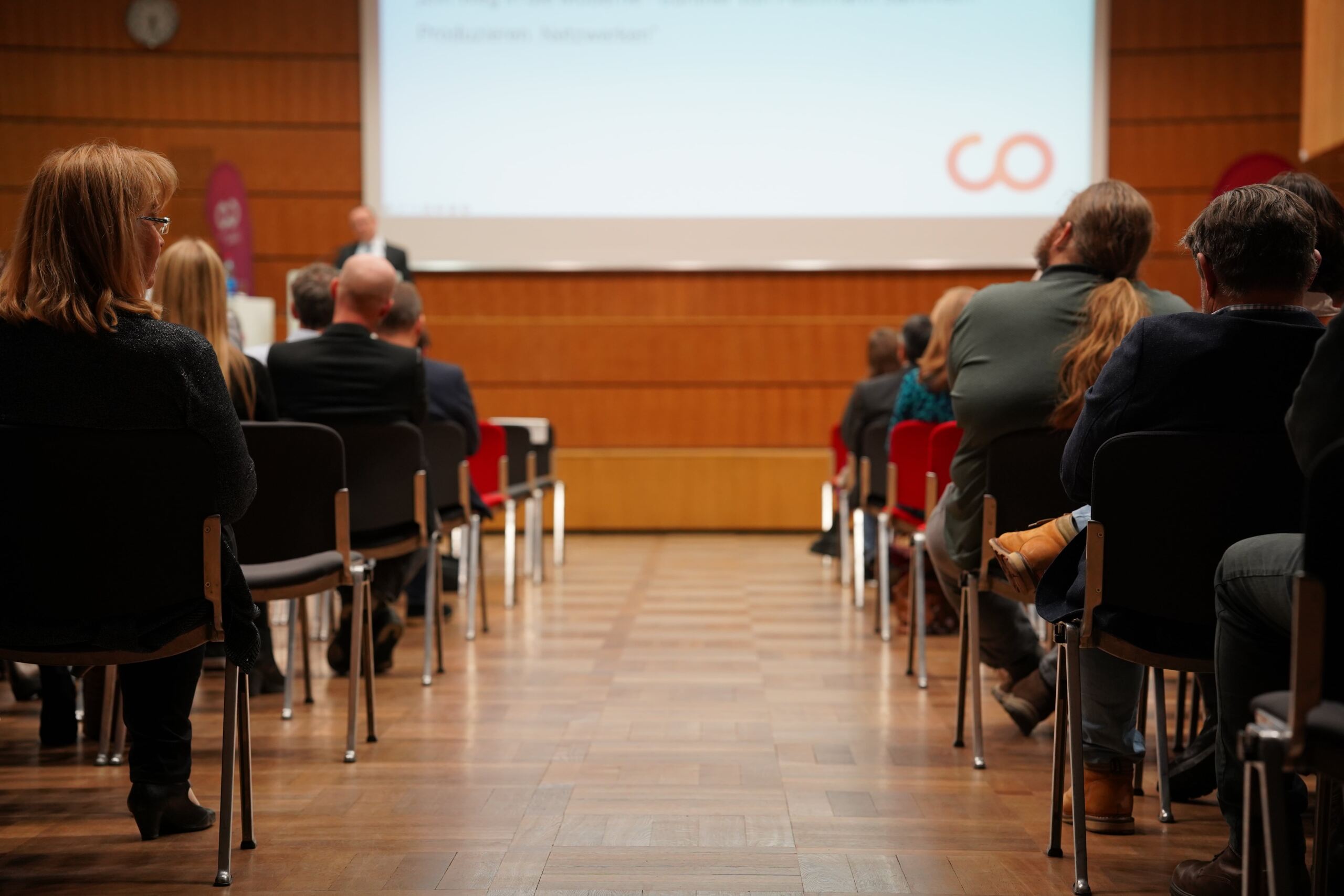 Das Publikum sitzt in einem Konferenzraum vor einer Holzbühne mit einem Vortragenden vor einem Bildschirm, auf dem eine Präsentation läuft. Stuhlreihen erstrecken sich nach vorn, und die Leute hören aufmerksam zu.