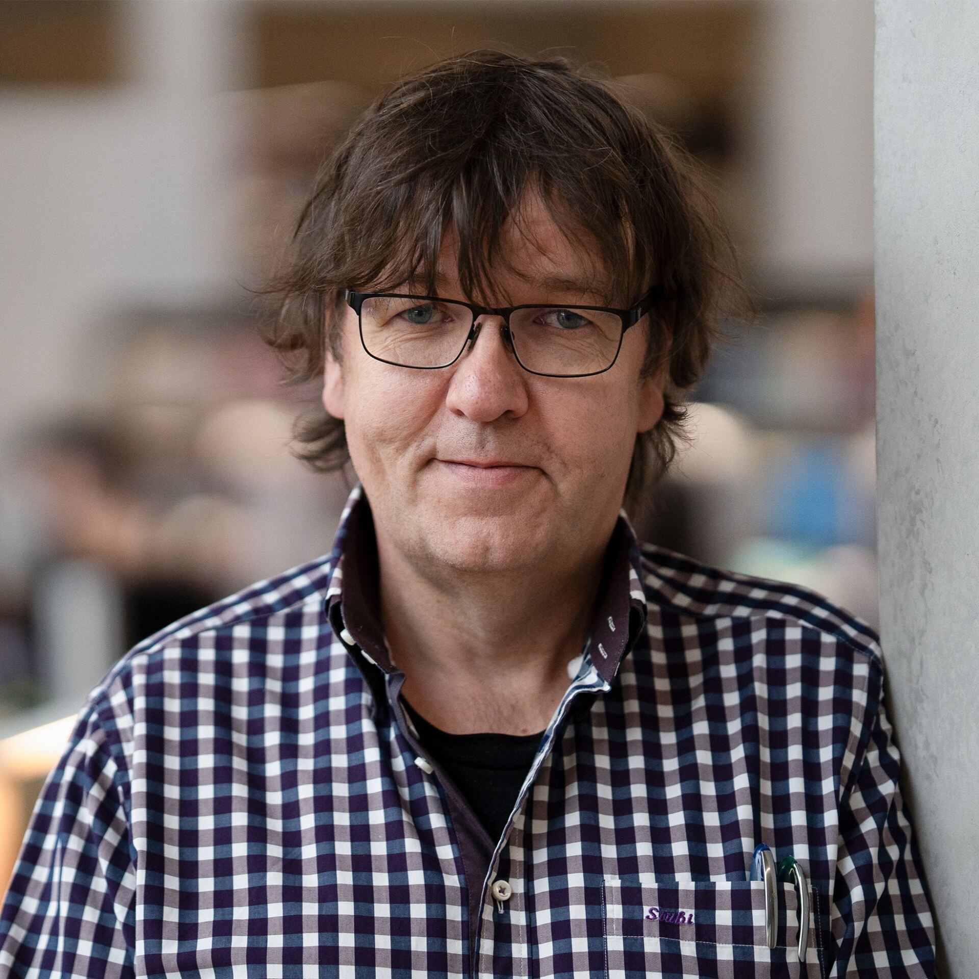 A person wearing glasses and a checkered shirt is standing indoors. They have medium-length hair and are looking directly at the camera. The background is softly blurred. at Hochschule Coburg