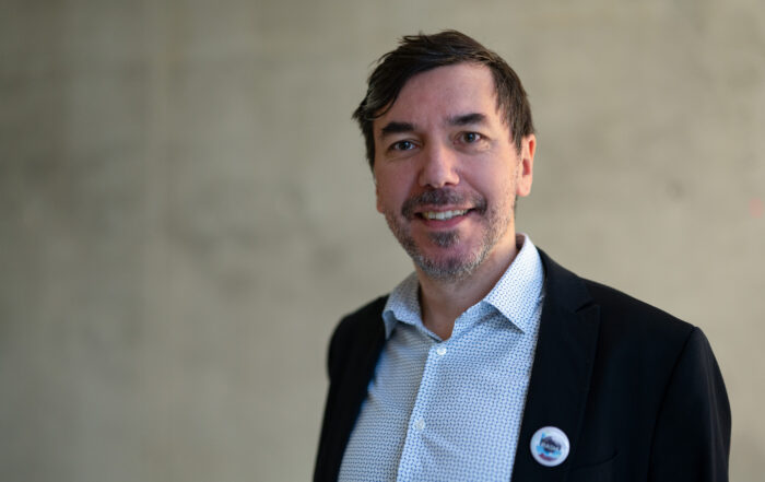 Ein Mann mit kurzen Haaren und Bart lächelt in die Kamera. Er trägt einen schwarzen Blazer über einem weißen gemusterten Hemd und auf seinem Blazer befindet sich ein kleines rundes Abzeichen. Der Hintergrund ist schlicht und verschwommen. at Hochschule Coburg