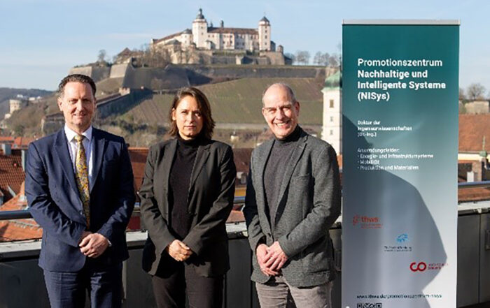 Drei Personen stehen auf einer Terrasse mit malerischer Aussicht auf ein historisches Schloss und einem klaren blauen Himmel im Hintergrund. Sie stehen neben einem Banner mit einem deutschen Text, der sich auf ein Promotionszentrum Nachhaltige Systeme (NiSyS) bezieht. at Hochschule Coburg