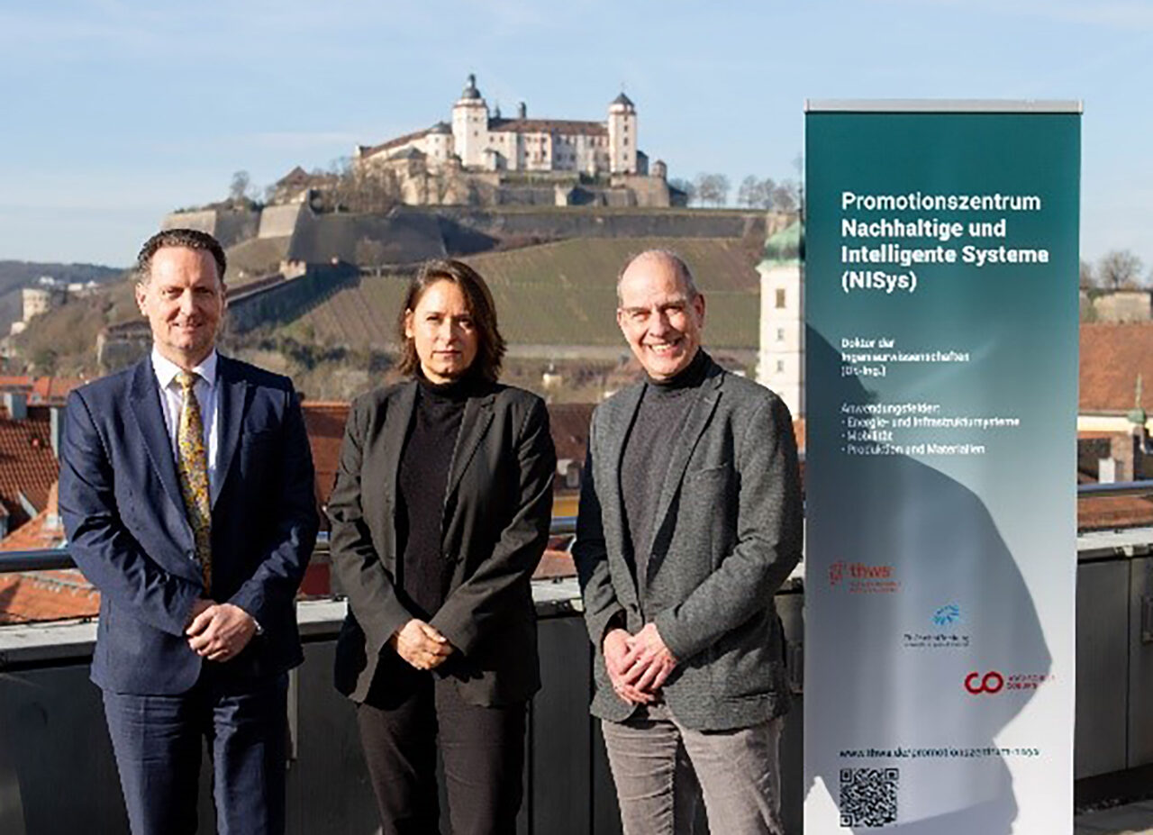 Drei Personen stehen auf einer Terrasse mit malerischer Aussicht auf ein historisches Schloss und einem klaren blauen Himmel im Hintergrund. Sie stehen neben einem Banner mit einem deutschen Text, der sich auf ein Promotionszentrum Nachhaltige Systeme (NiSyS) bezieht. at Hochschule Coburg