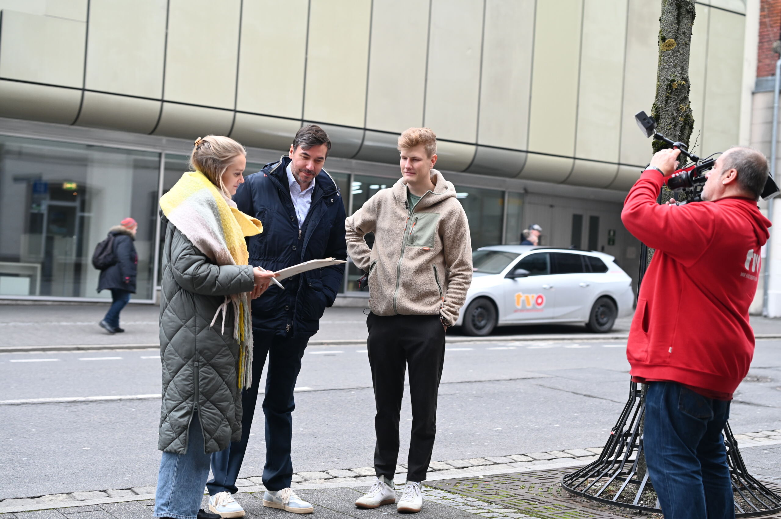 Eine Frau, ein Mann und ein junger Mann stehen auf einem Bürgersteig neben einem weißen Auto. Die Frau hält einen Stapel Unterlagen zu ihrem Architekturstudium in der Hand. In der Nähe filmt sie jemand in einer roten Jacke mit einer großen TVO-Kamera, während im Hintergrund ein Gebäude mit großen Fenstern aufragt. at Hochschule Coburg