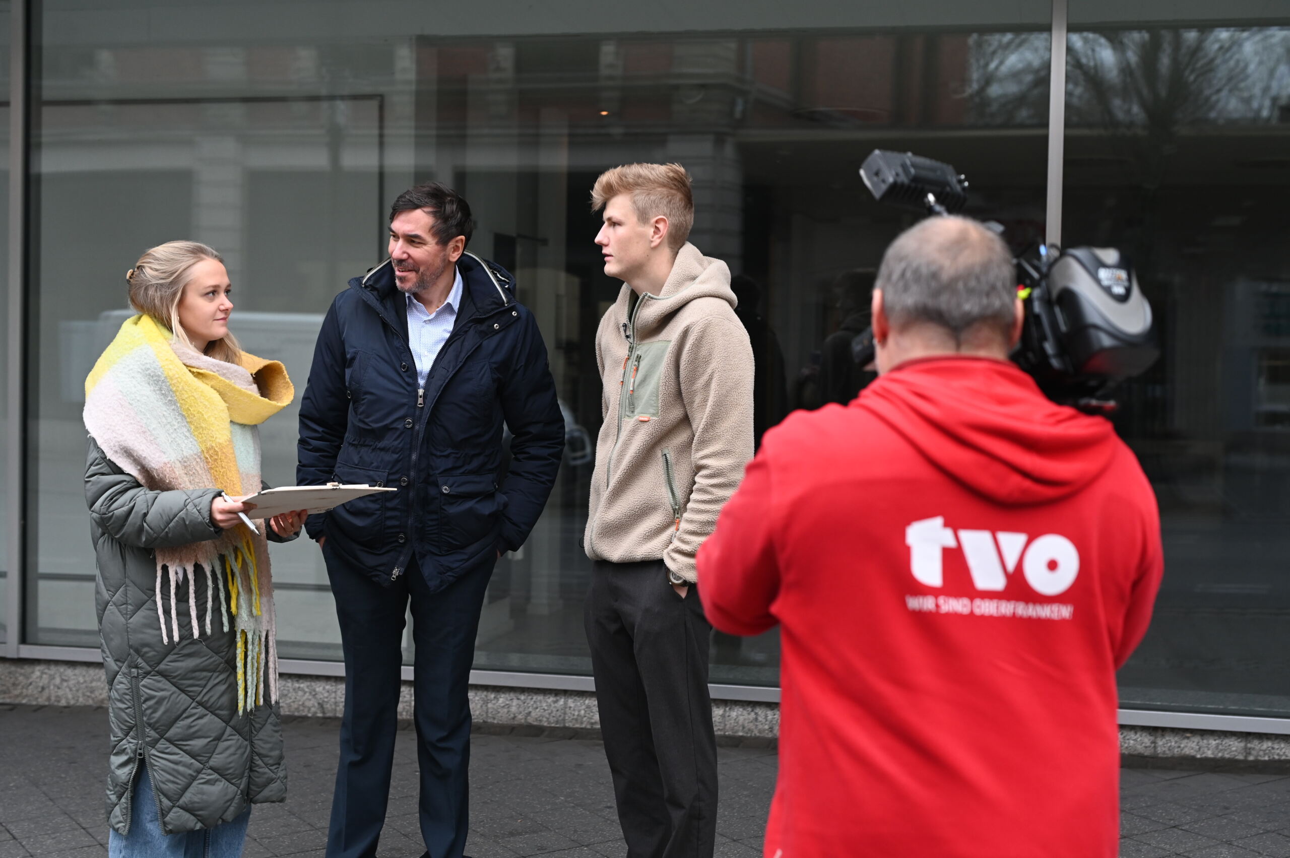 Eine Frau und zwei Männer unterhalten sich auf dem Bürgersteig. Die Frau hält ein Klemmbrett in der Hand. In der Nähe filmt sie jemand in einer roten Jacke mit der Aufschrift „TVO“ auf dem Rücken vor dem Hintergrund eines großen Fensters, das an den Architekturstil des Kaufhofs erinnert. at Hochschule Coburg