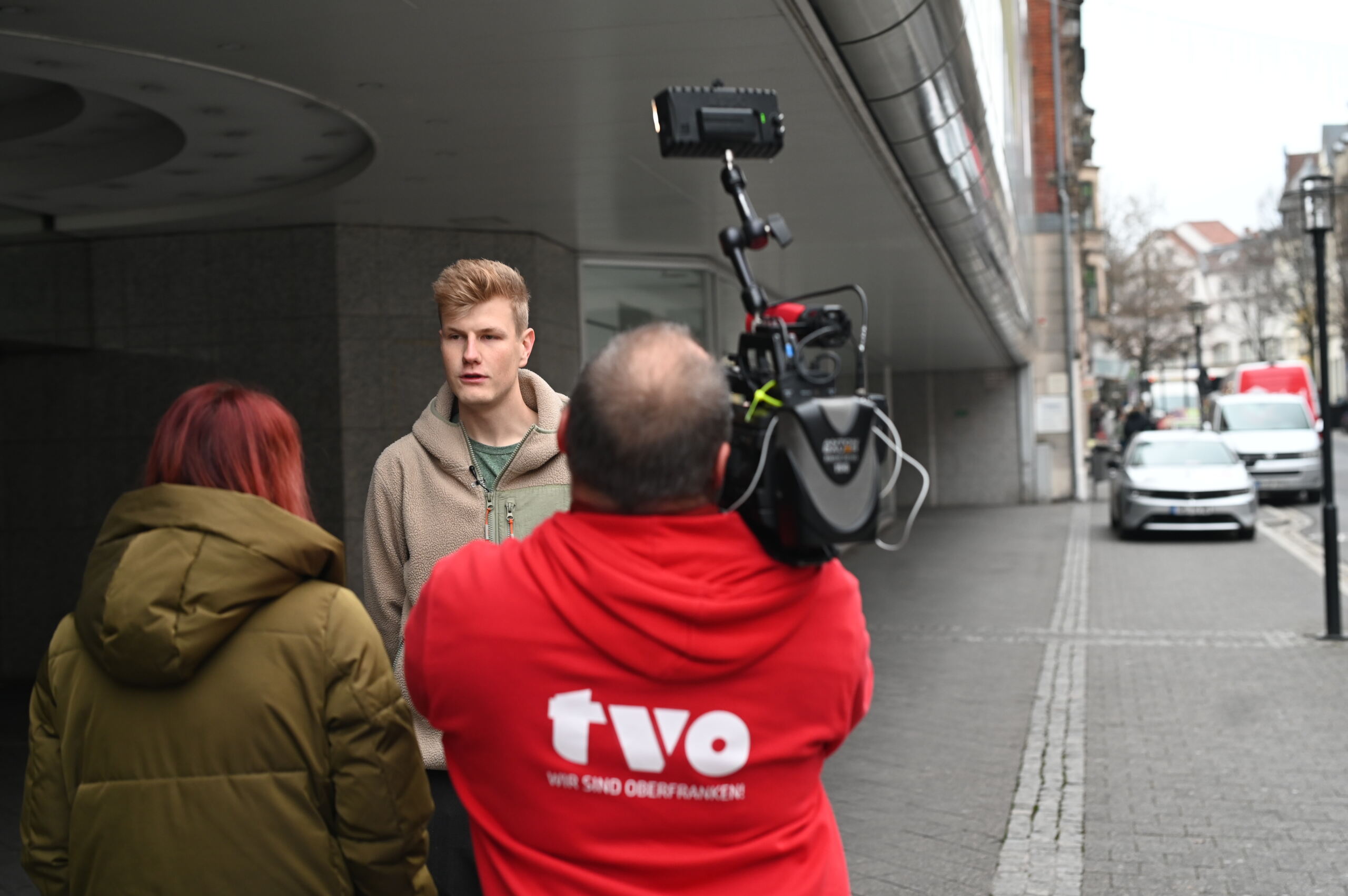 Eine Person mit einer Kamera auf der Straße interviewt einen Mann in einem hellbraunen Kapuzenpullover, während ein anderer in einer grünen Jacke zusieht. Ein weißer Lieferwagen steht in der Nähe im Schatten der Kaufhof-Architektur, und die Straße ist von Gebäuden gesäumt. Der Kameramann trägt eine rote Jacke mit der Aufschrift „TVO“ auf dem Rücken. at Hochschule Coburg